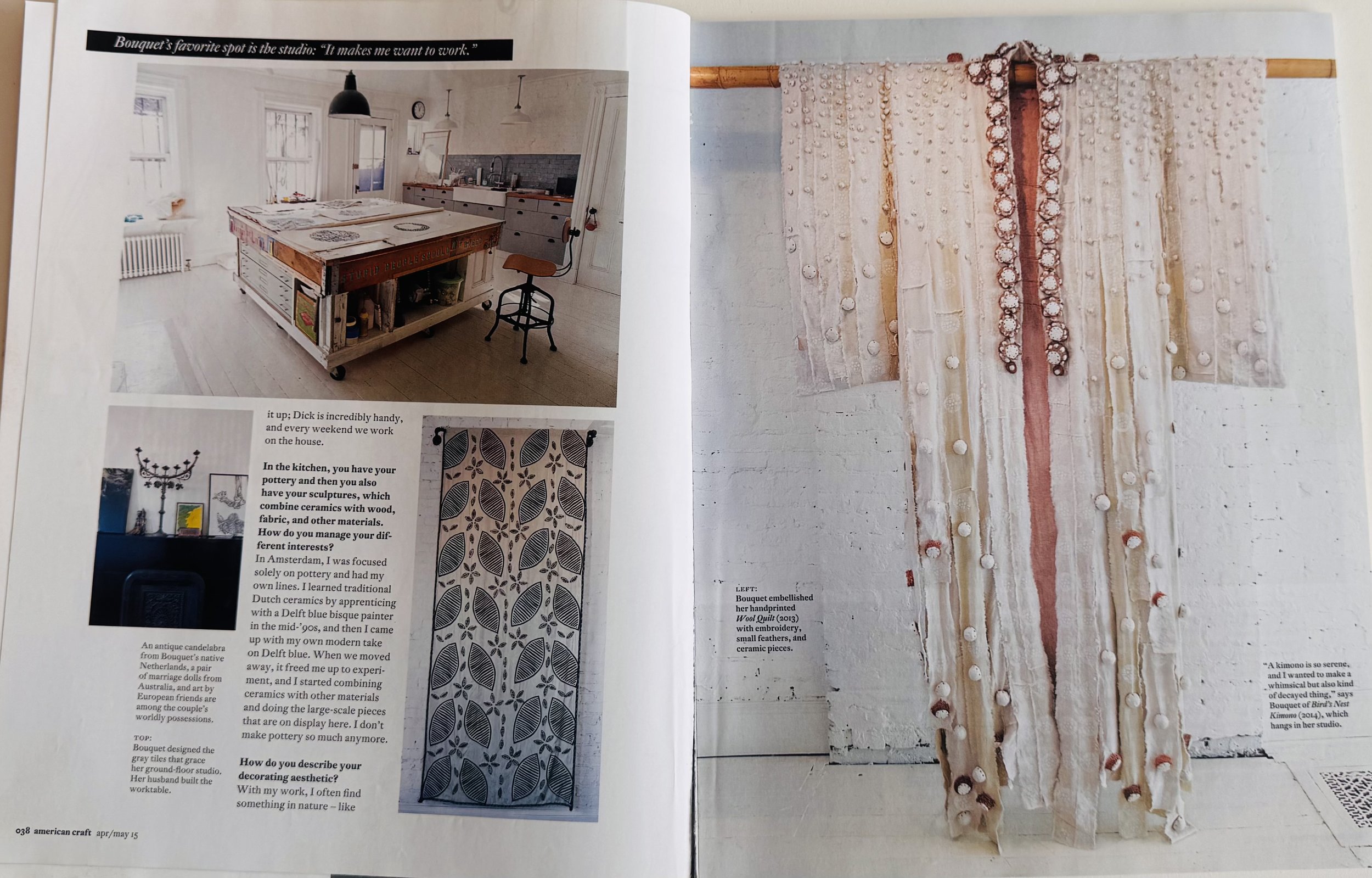 A two-page magazine spread featuring interior design and craft projects. The left page shows a modern kitchen with a large island, a stool, and decorative items on shelves, with a caption about working in the studio. The right page displays a fabric wall hanging with embroidered elements, described as a hand-printed wool quilt with embroidery, small feathers, and ceramic pieces, hanging on a bamboo rod against a white brick wall.