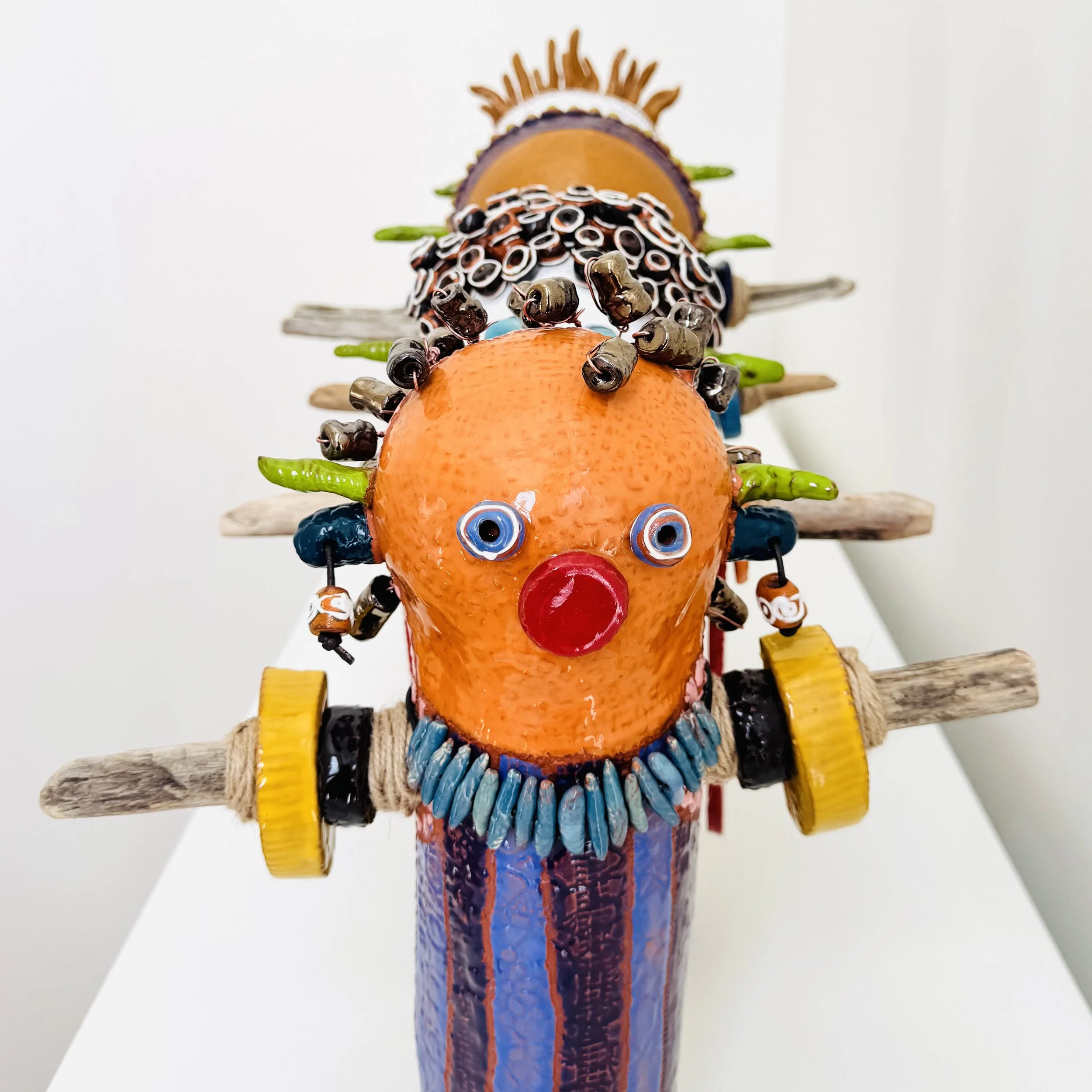 Colorful sculpture of a clown with an orange head, large red nose, wide blue eyes, wearing a necklace of blue stones, holding a yellow barbell with wooden sticks attached.