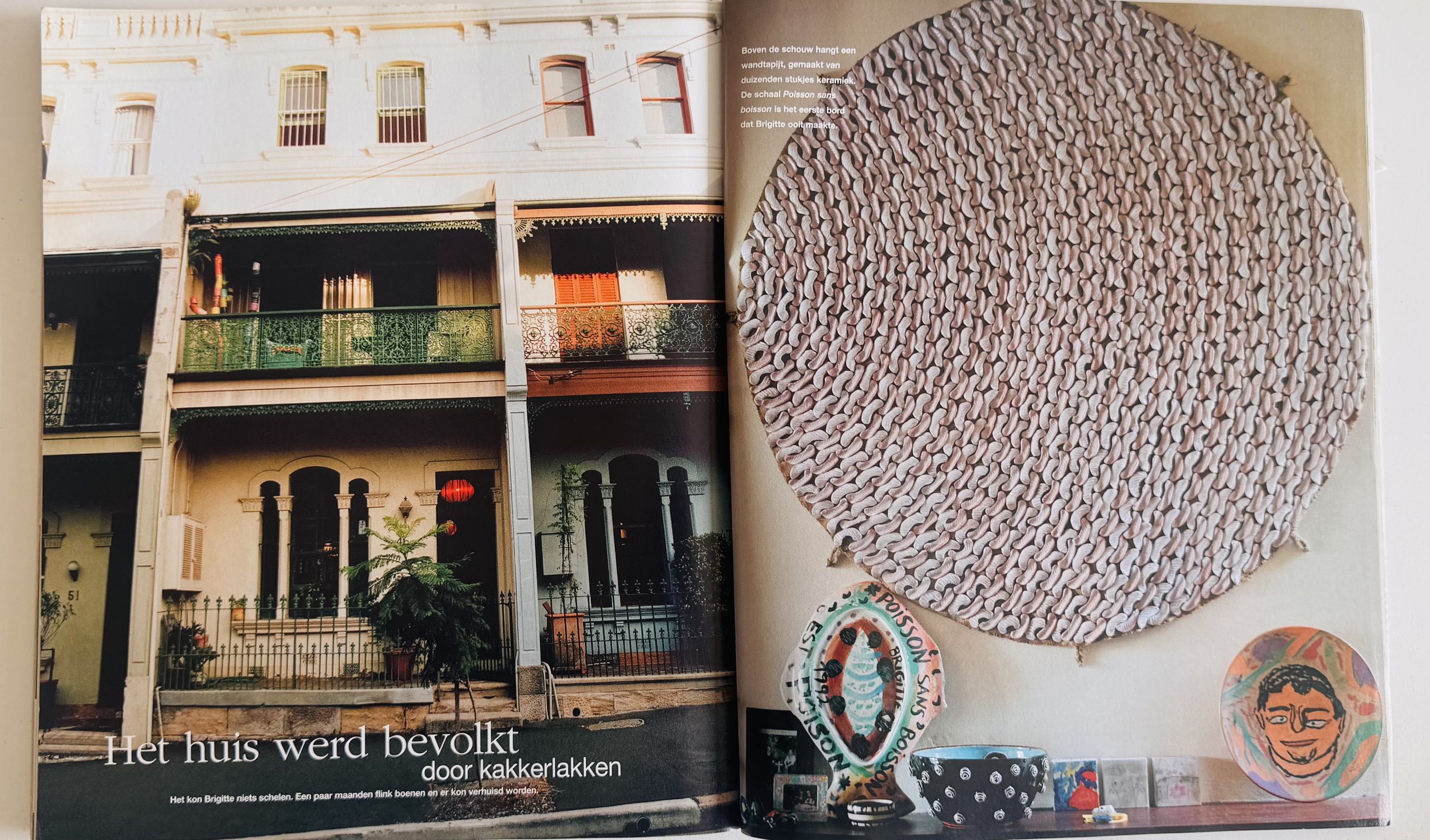 A two-page magazine spread. The left page shows a photograph of a white multi-story house with balconies, green and red railings, and decorative window frames. The right page features a large, circular woven placemat or rug made of pinkish-brown material, mounted on a wall with some artwork arranged below it, including drawings and a decorated plate.