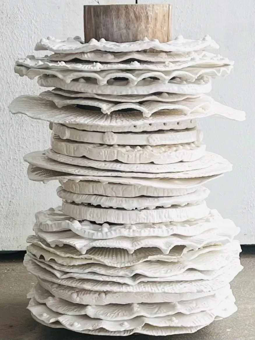 Stack of white ceramic or plaster flower-like sculptures placed on a concrete floor against a white wall, with a cylindrical wooden piece on top.