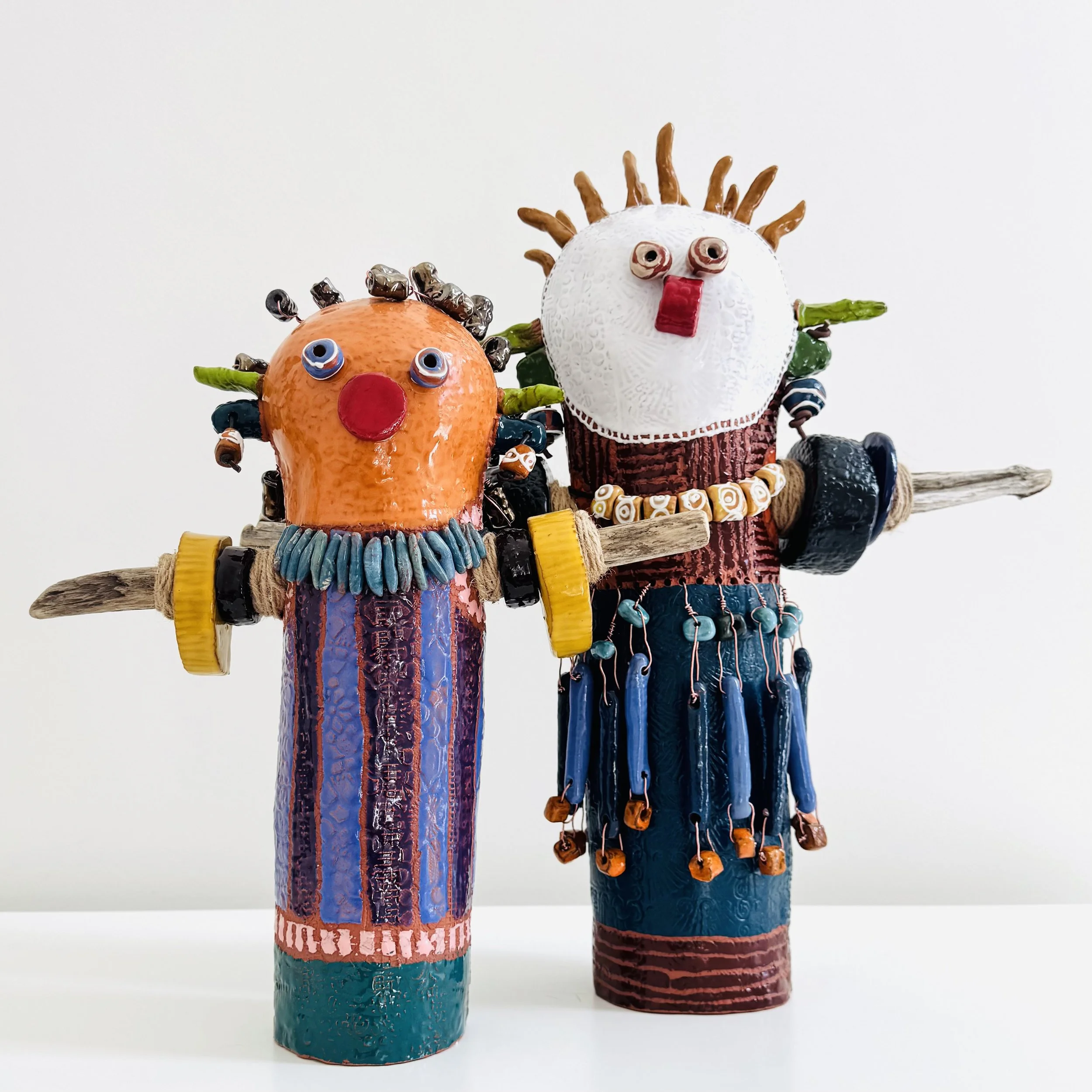 Two ceramic figures from the GOBI series by Brigitte Bouquet, each approximately eighteen inches tall. Vivid and richly textured, one figure with an orange ceramic head, one with white, their arms formed from driftwood sticks
