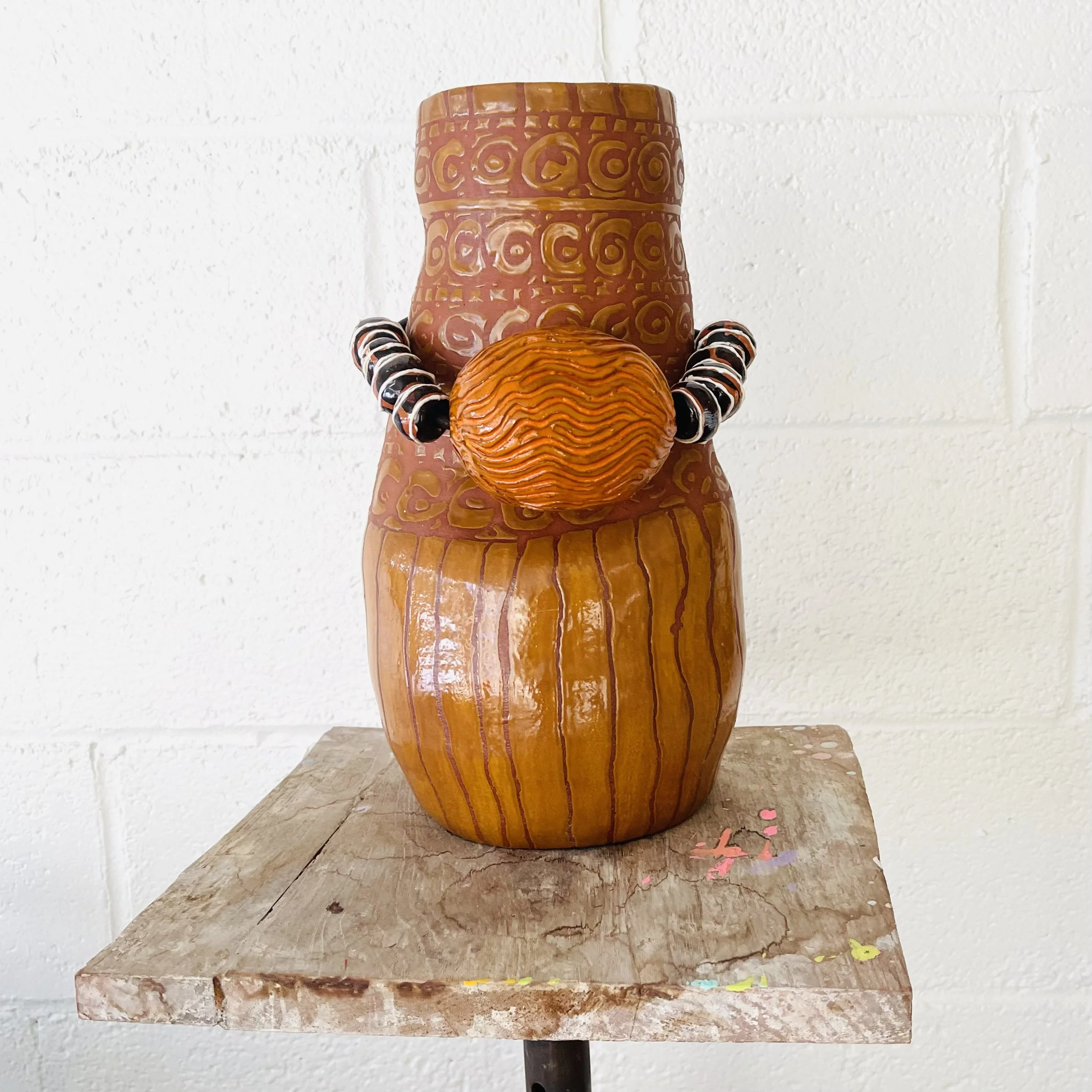 Decorative ceramic sculpture of a human figure with an orange body, large round textured nose, wearing black and white bead necklaces, placed on a wooden stand against a white brick wall.