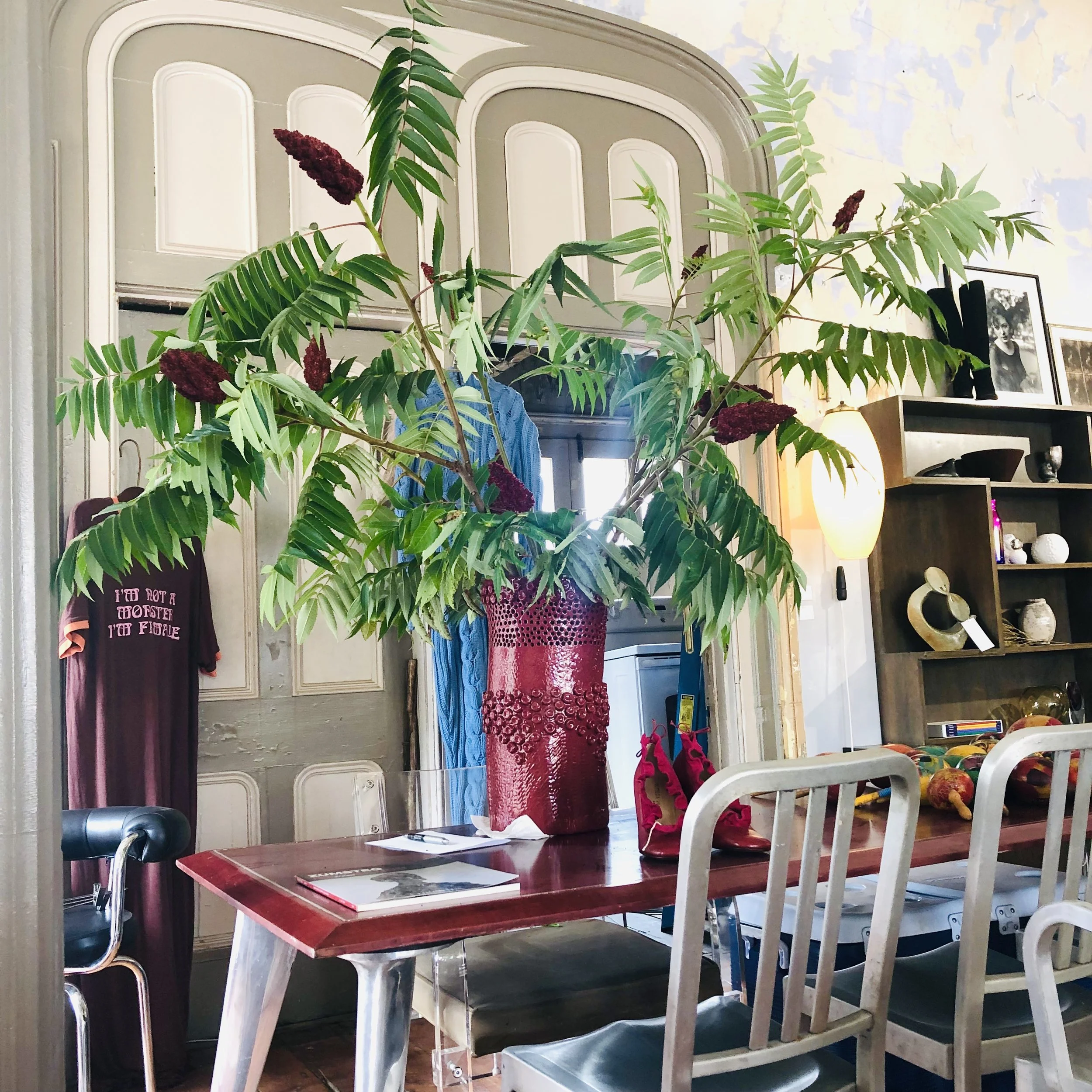 A large potted plant with green leaves and purple flowers on a red vase is placed on a wooden table in a room with bookshelves, framed pictures, and a wall lamp.