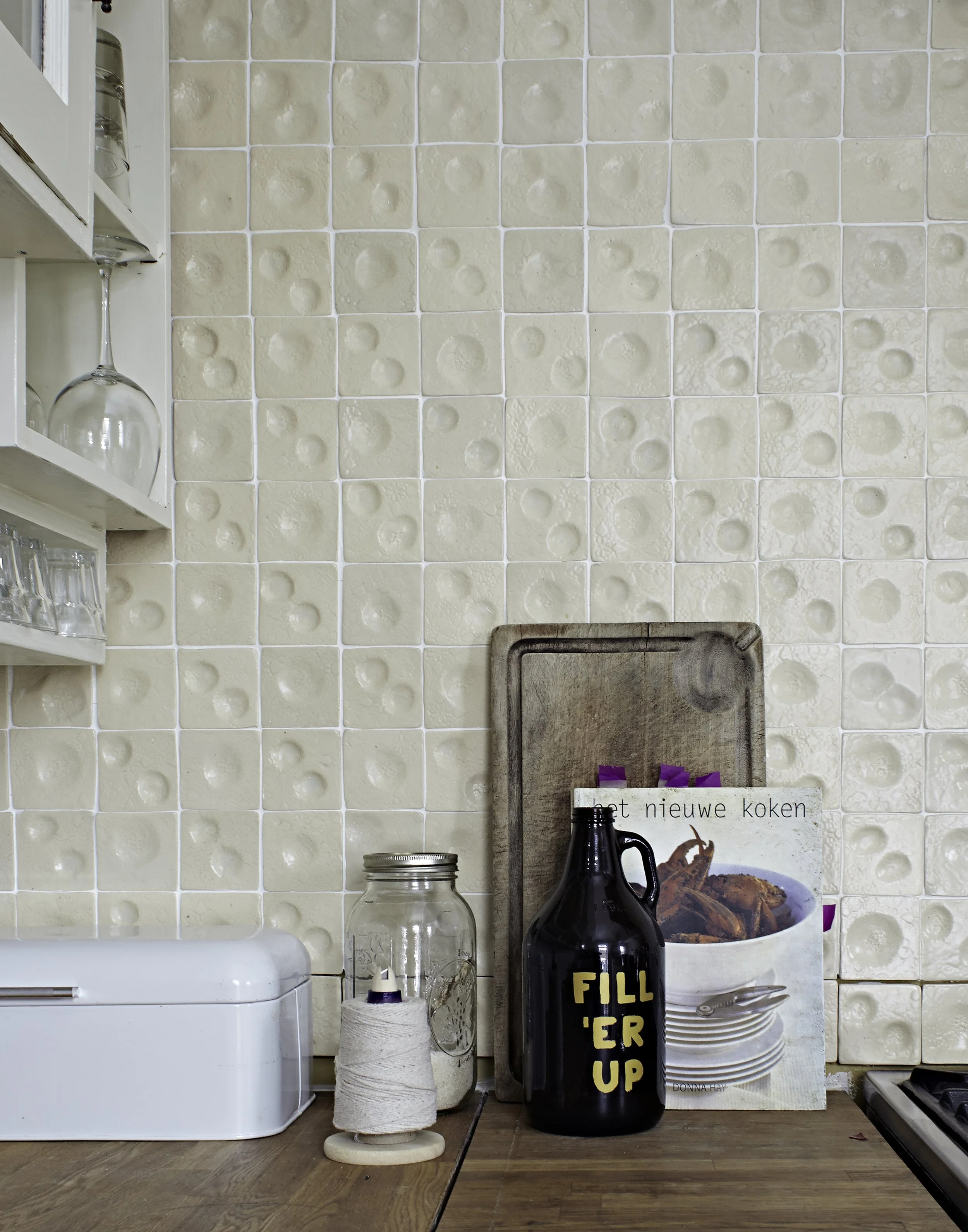 A kitchen countertop with a white container, a glass jar, a black bottle with yellow text reading 'FILL 'ER UP', a wooden cutting board, a cookbook, and a spool of twine. Patterned beige tile backsplash with cups and glasses on shelves.