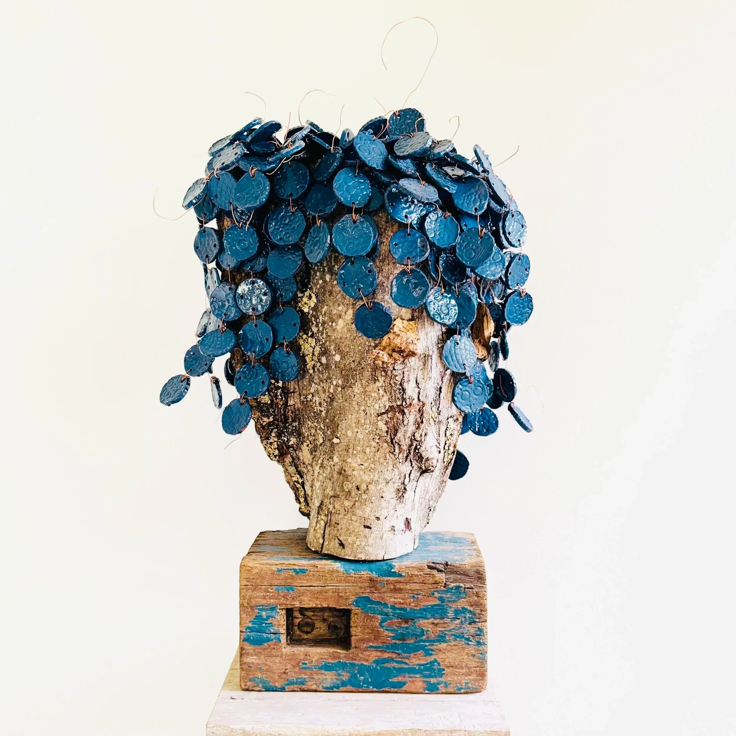 A sculpture of a face with blue circular leaves or discs for hair, mounted on a rustic wooden base.