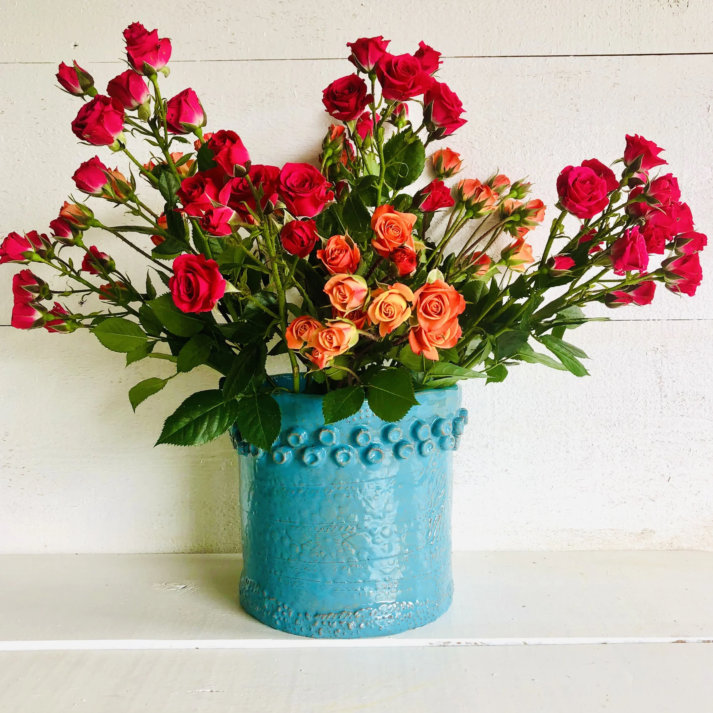 A blue ceramic vase filled with pink and orange roses, placed on a white surface against a white wall.