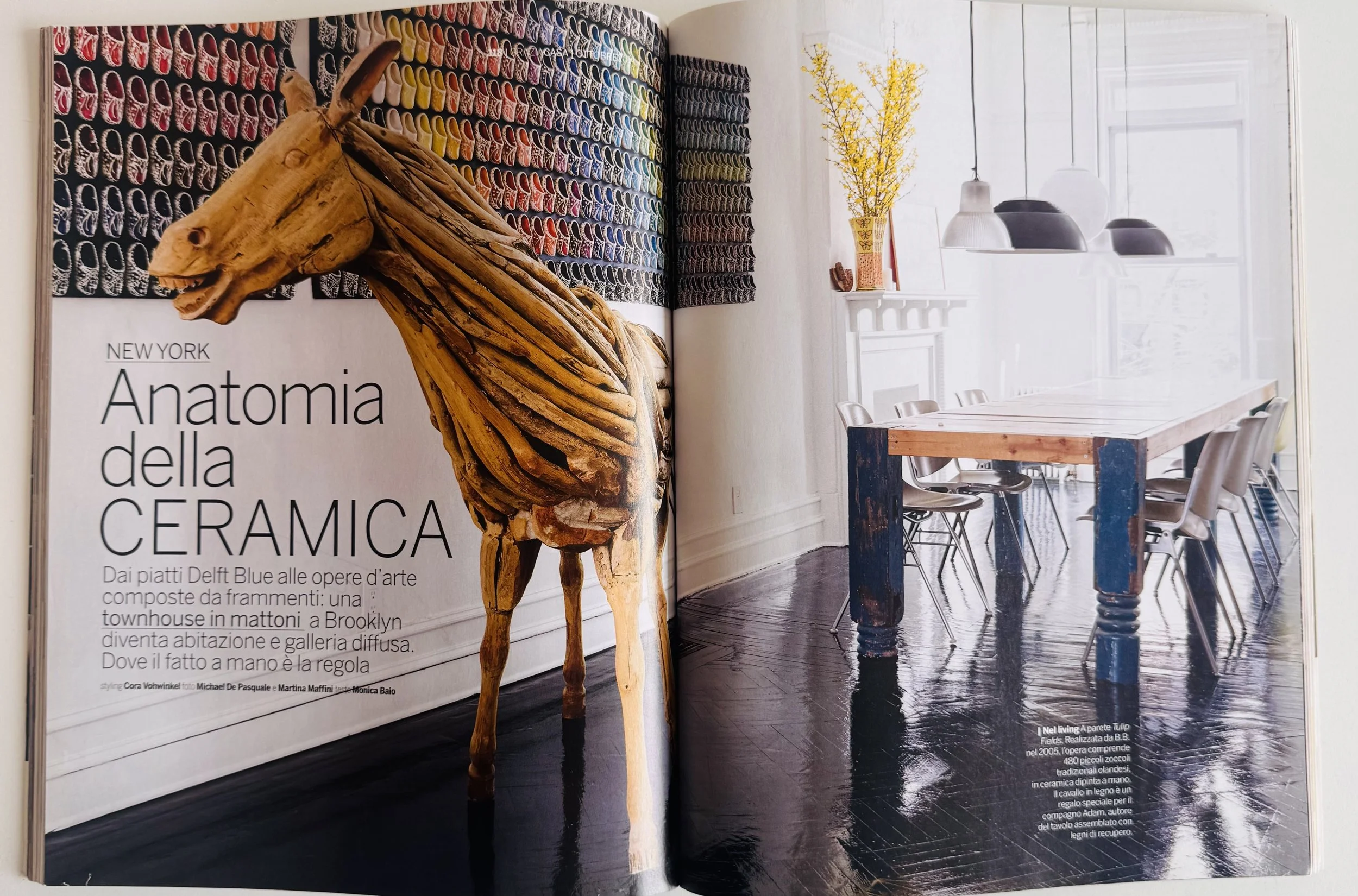 Open magazine showing a room with a large wooden dining table, white chairs, a window with natural light, decorative lamps, a white fireplace mantel with a yellow flowering plant, and a large wall art of a shoe display and a wooden horse sculpture.