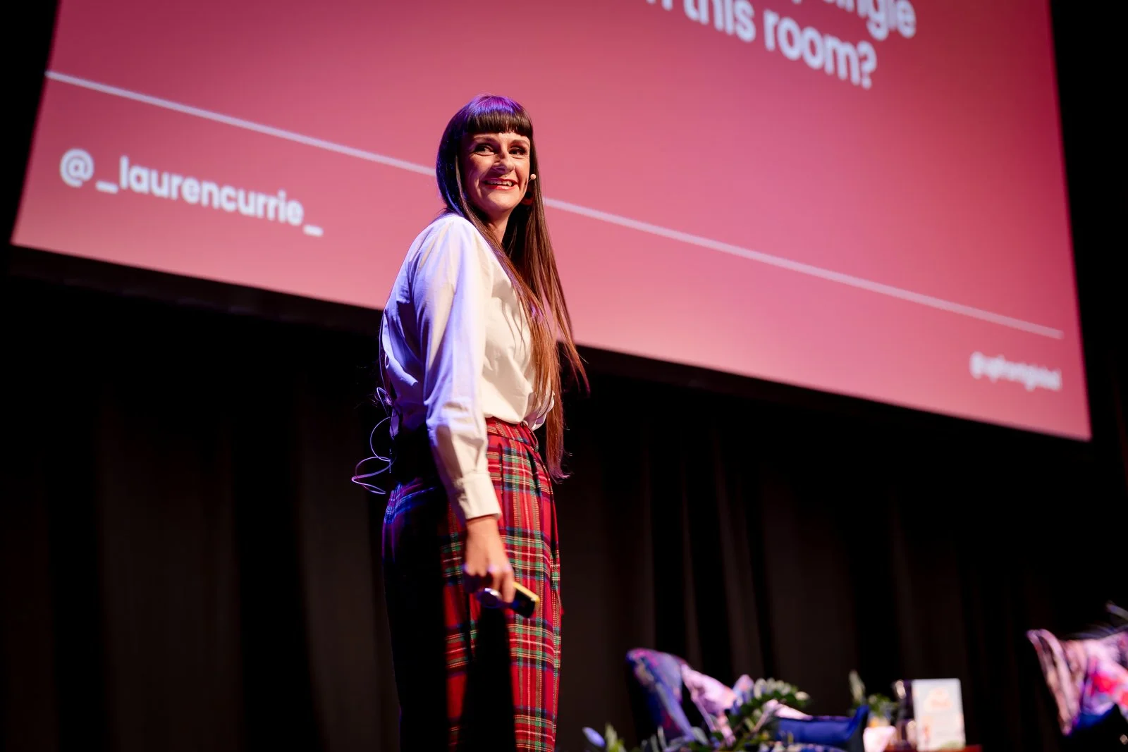 A woman with long brown hair and bangs, wearing a white shirt and red plaid pants, stands on stage holding a microphone, smiling. Behind her is a large screen with pink backdrop and white text, including an Instagram handle @_laurencurrie_.