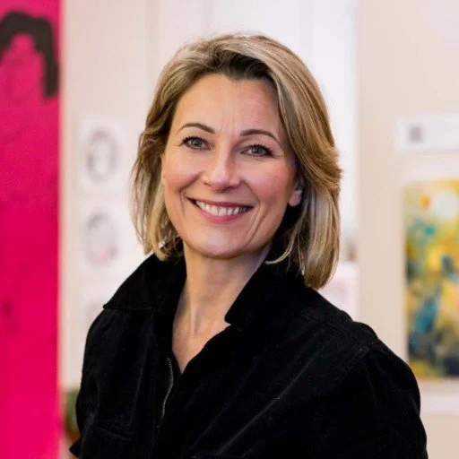 A smiling woman with shoulder-length blonde hair posing indoors, wearing a black top.