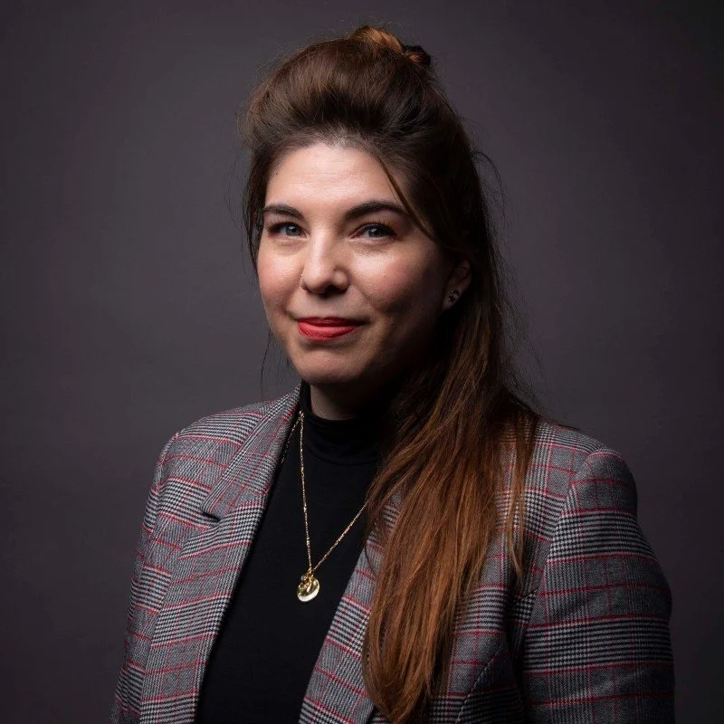 A woman with long brown hair styled in a tea-length puff, wearing a gray checkered blazer over a black top, gold pendant necklace, and small earrings, posing against a dark gray background.