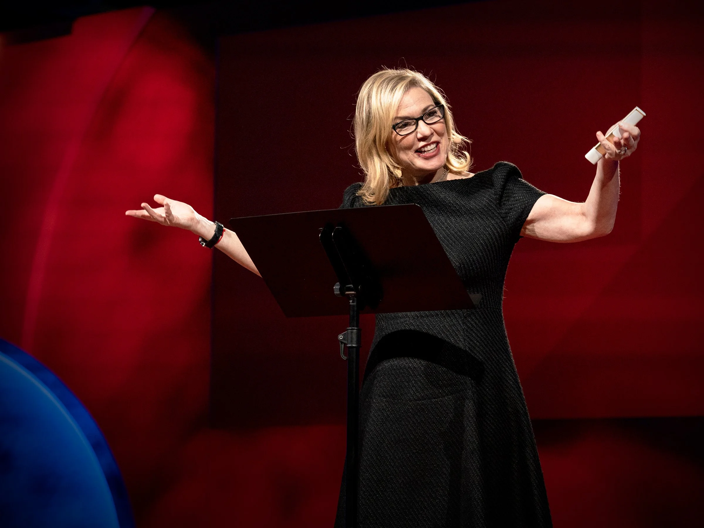 A woman with blonde hair and glasses, smiling and gesturing with her arms while speaking on stage.