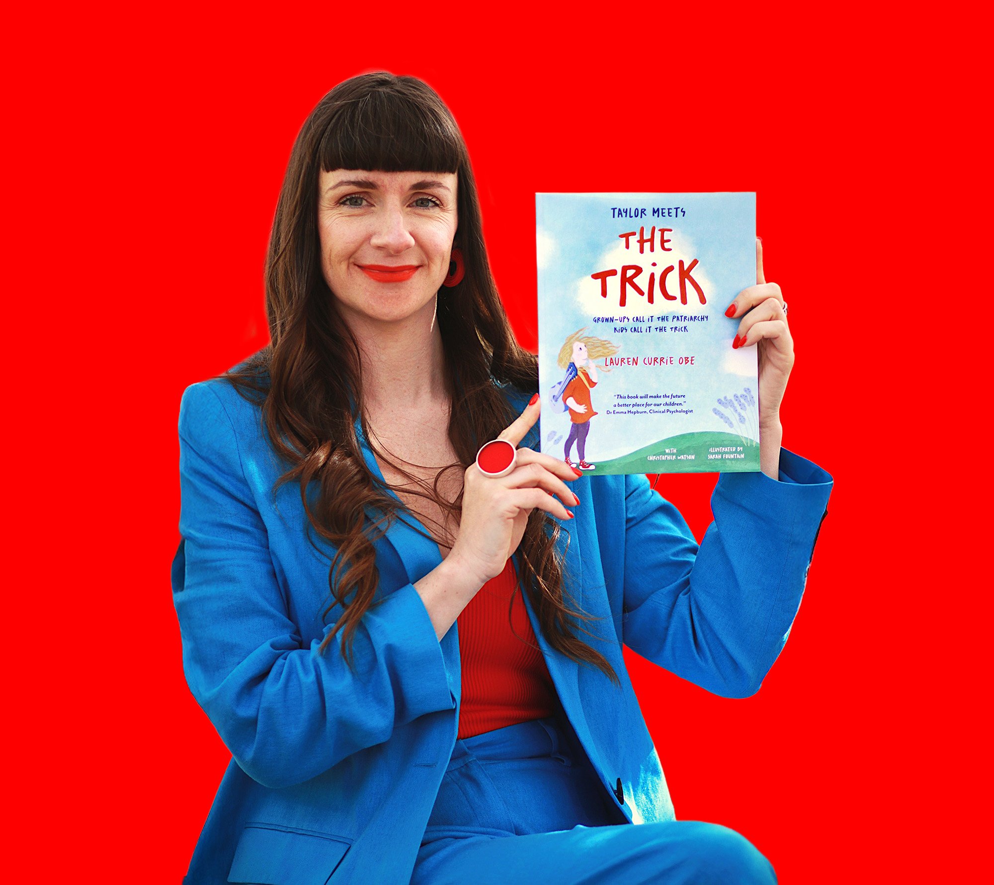A woman with long brown hair and red lipstick, dressed in a blue blazer, holding up a book titled "The Trick" by Lauren Currie Obe, against a bright red background.