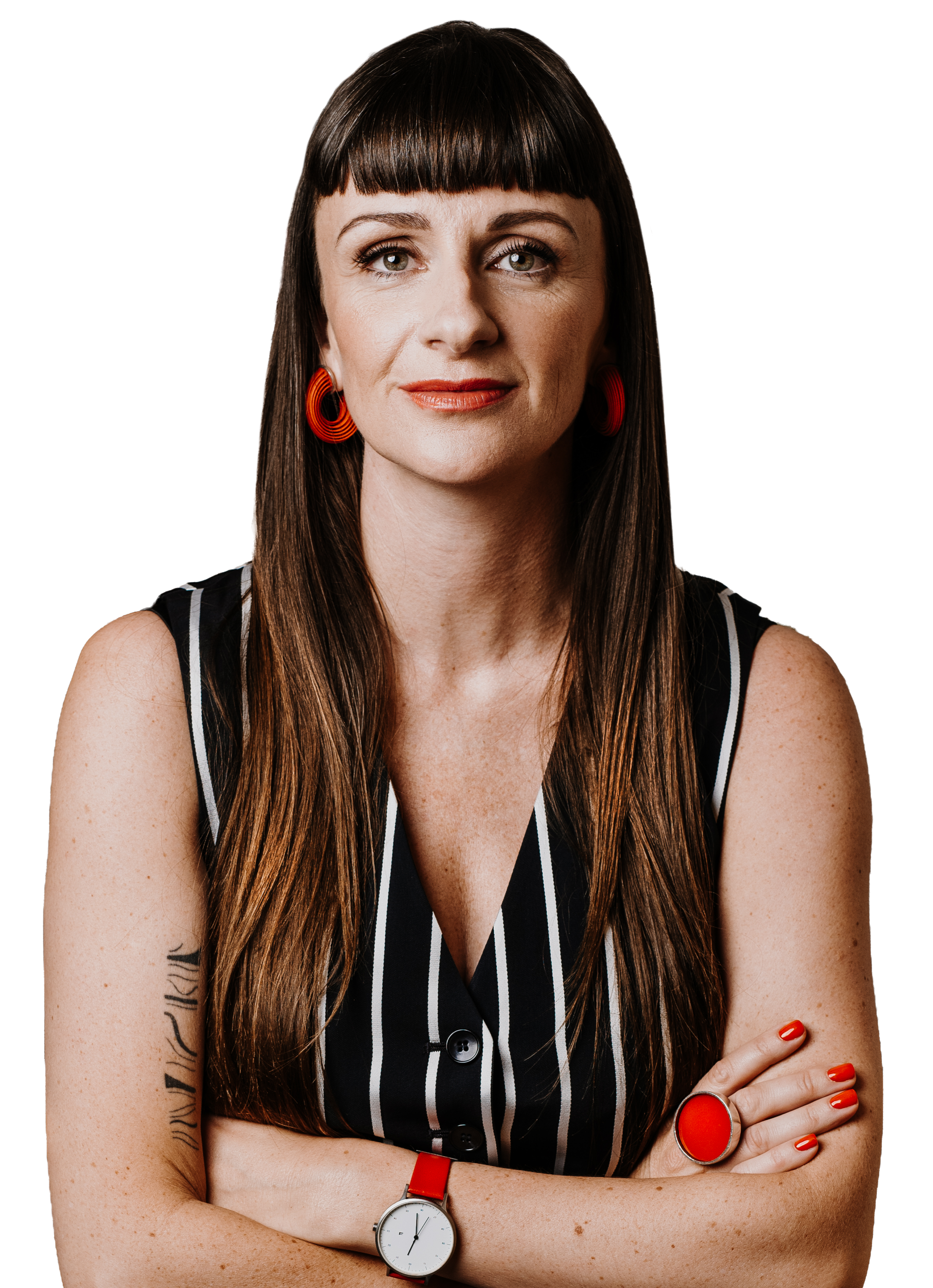 A woman with long brown hair and bangs, wearing a black and white striped sleeveless top, orange earrings, a large red ring, a red bracelet, and a white wristwatch with a red strap, standing with her arms crossed against a black background.