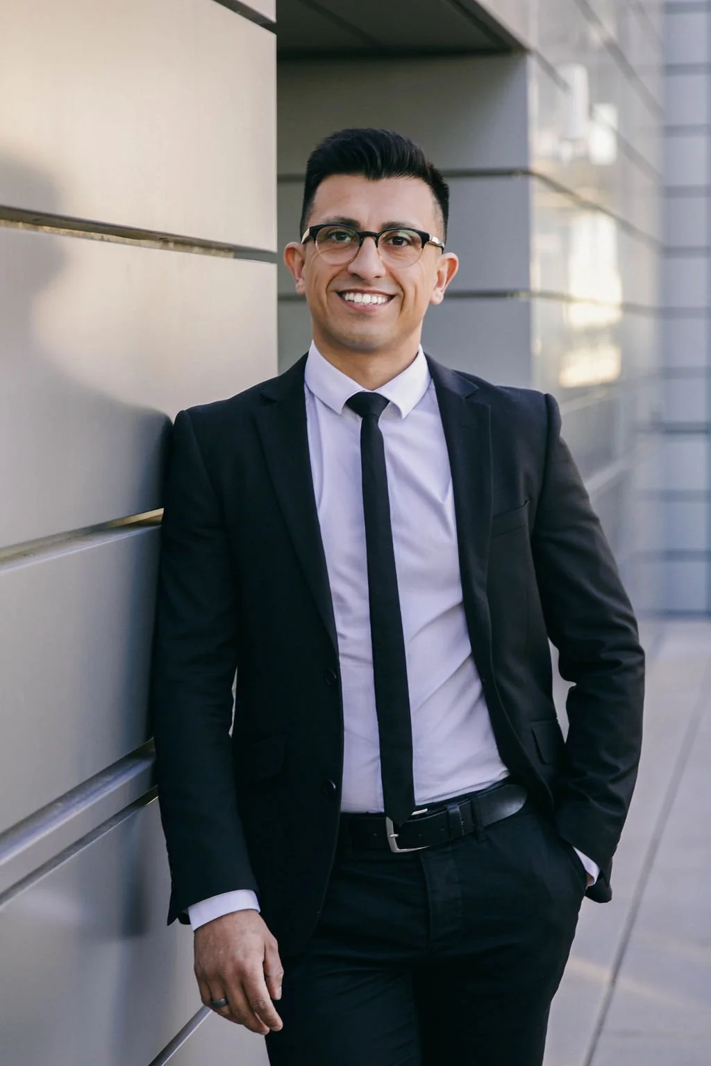 A smiling man in a black suit, white shirt, and black tie, wearing glasses, standing outdoors next to a modern building wall with glass windows.