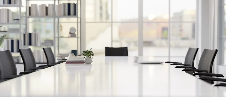 Empty conference room with a long table, chairs, a small plant, books, a coffee cup, and large windows in the background.