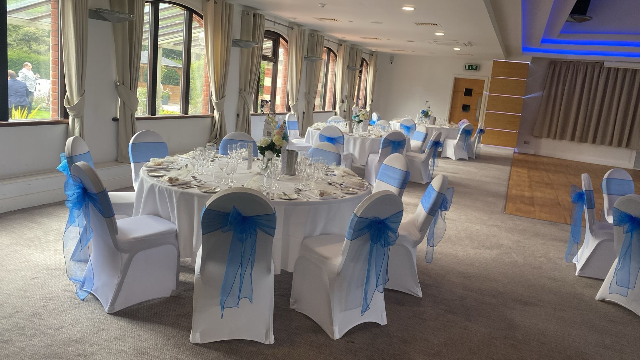 Wedding reception setup with round tables covered in white tablecloths, decorated with blue sashes on the chairs, floral centerpieces, and table settings including glasses and silverware, inside a room with large windows and curtains.
