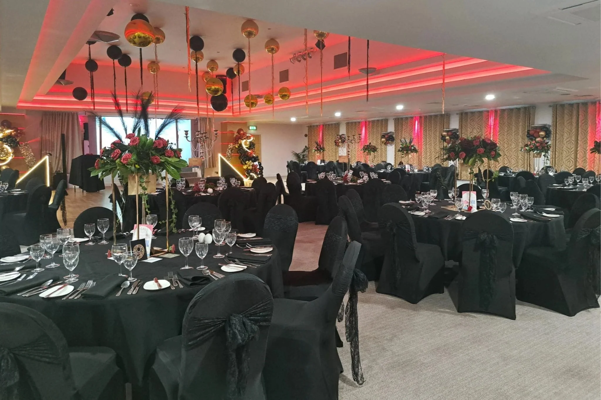A banquet hall decorated with black tablecloths, floral centerpieces, and hanging ornaments with gold and black accents, illuminated with pink and red lighting.