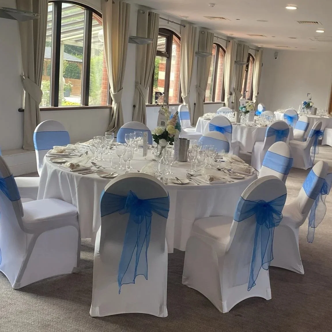 Interior of a banquet hall decorated with white tablecloths and white chairs adorned with blue sashes, set with glasses, plates, and utensils, and decorated with floral centerpieces, with large windows and cream curtains.