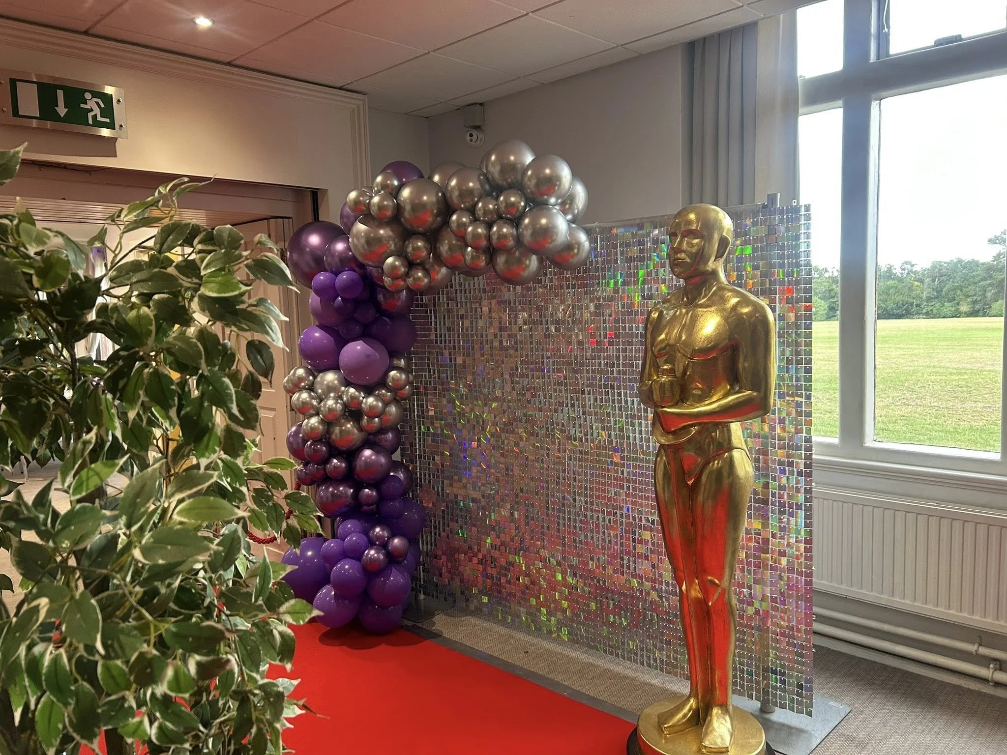 Gold mannequin sculpture of a woman with a short hairstyle, standing on a circular base. Behind the sculpture, a colorful holographic mosaic wall. To the left, a balloon arch with purple, silver, and metallic balloons. A red carpet in front of the di