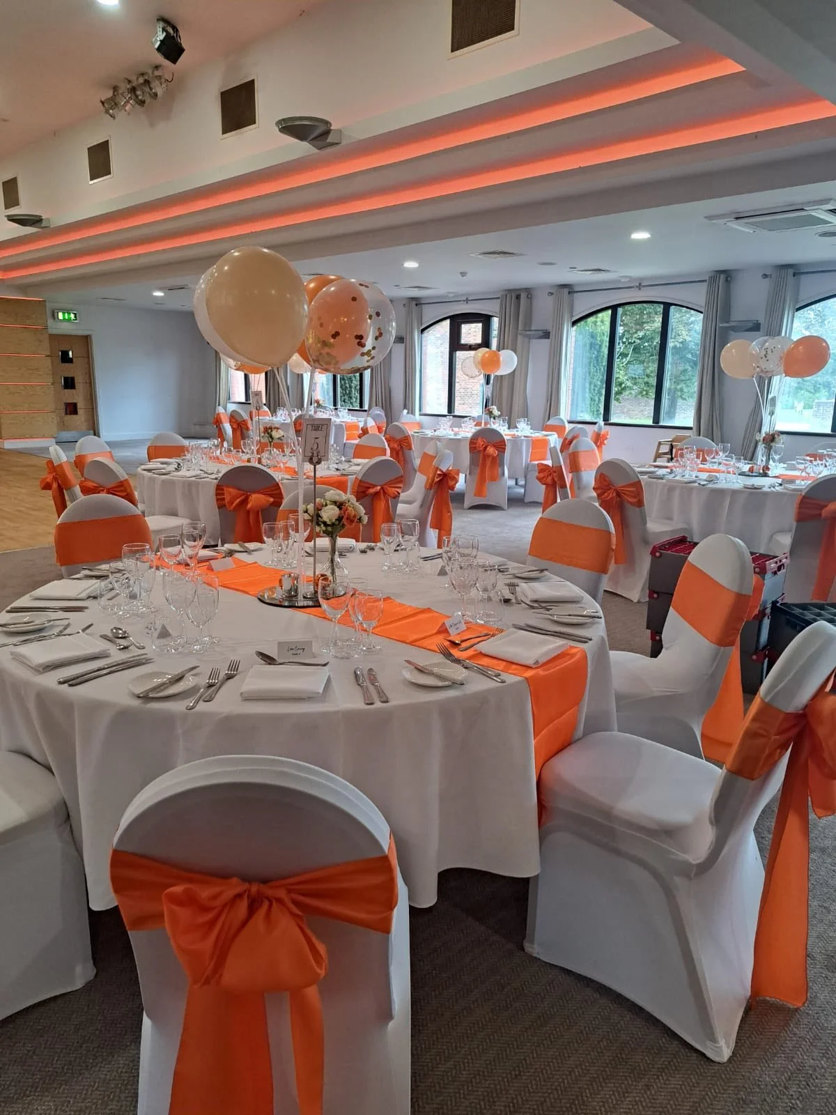 Set up banquet hall with round tables covered in white tablecloths, decorated with orange ribbons on the chairs, and centerpieces of flowers and balloons. Large windows let in natural light.