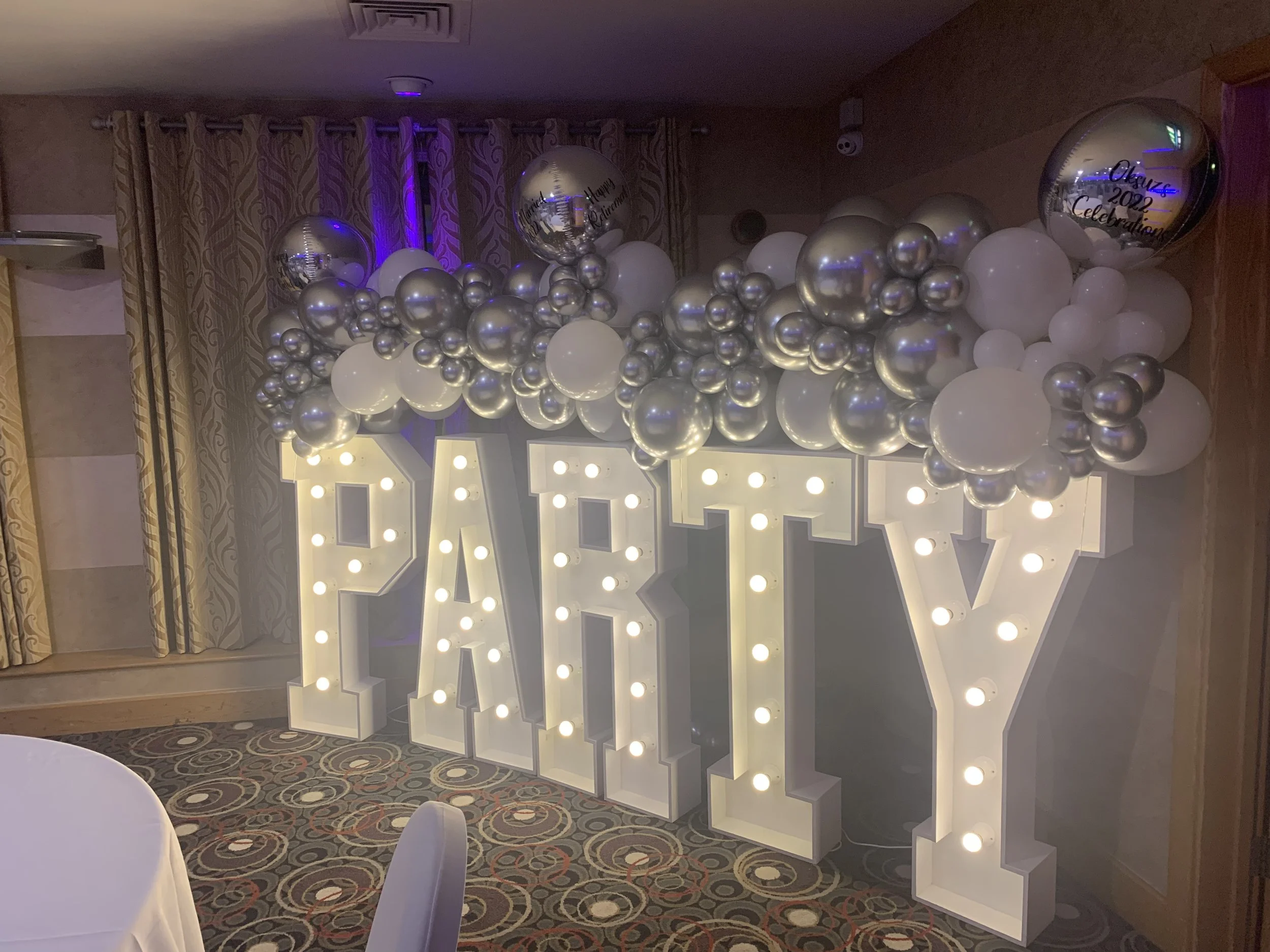 Decorative wedding display with large illuminated letters spelling 'PARTY' surrounded by white and silver balloons, some with message text, in an event space with curtains and patterned carpet.