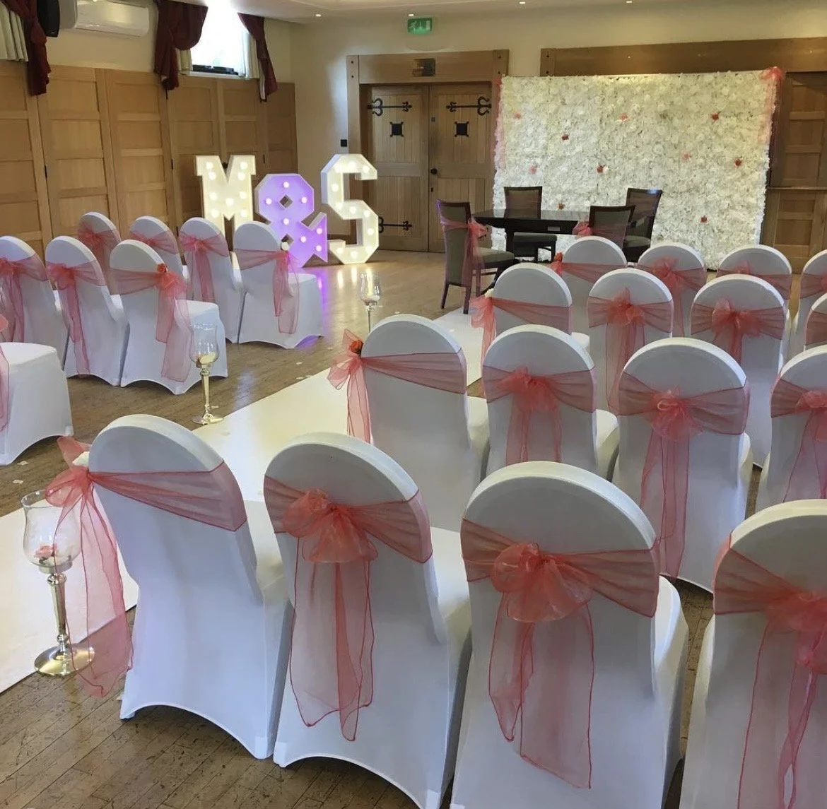 Wedding or event reception setup with white chairs decorated with pink sheer sashes, and a large illuminated decoration displaying the letters 'M & S'.