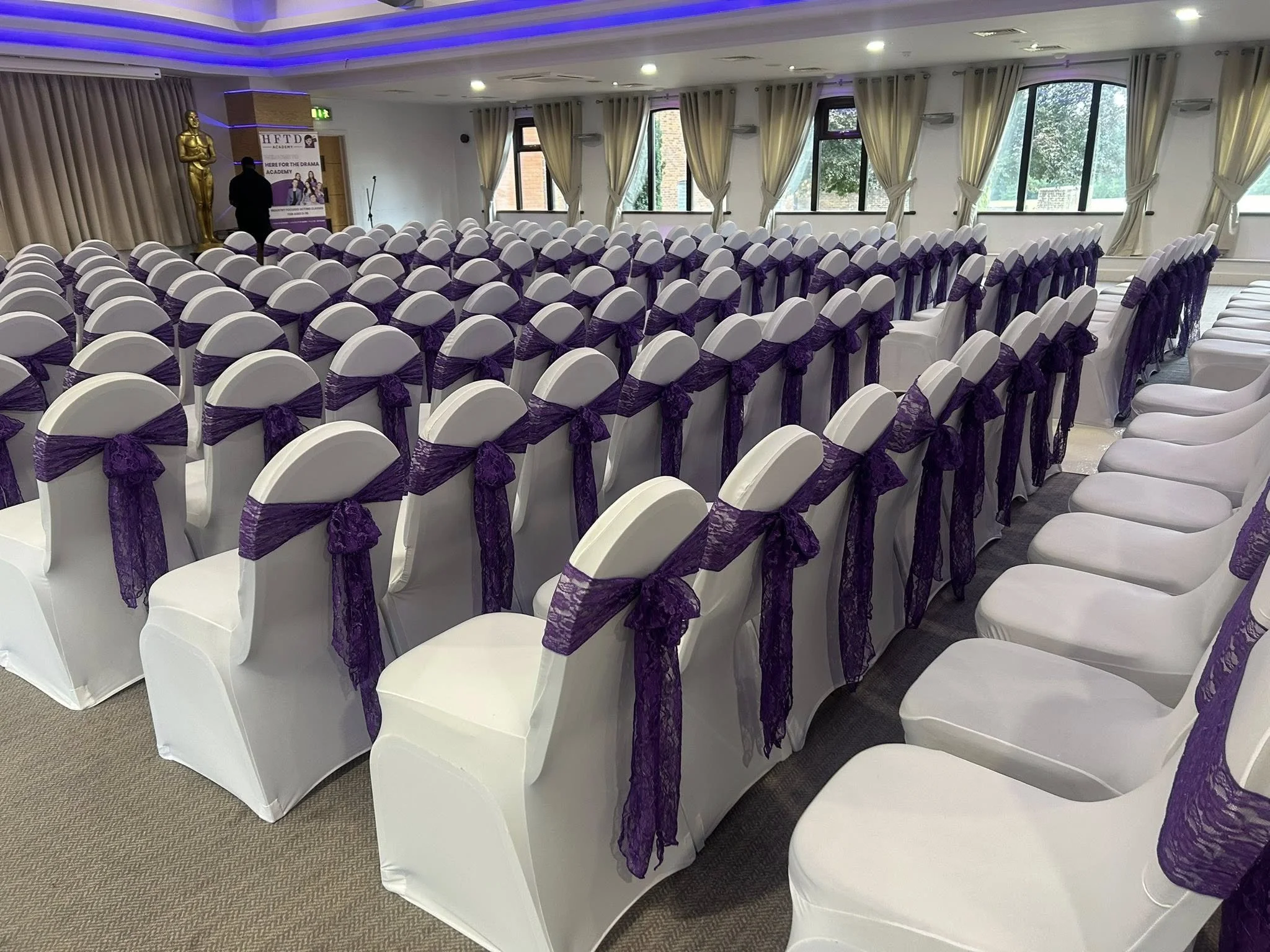 A room set up for a formal event with rows of white chairs decorated with purple sashes, facing a stage area with a statue and a banner.
