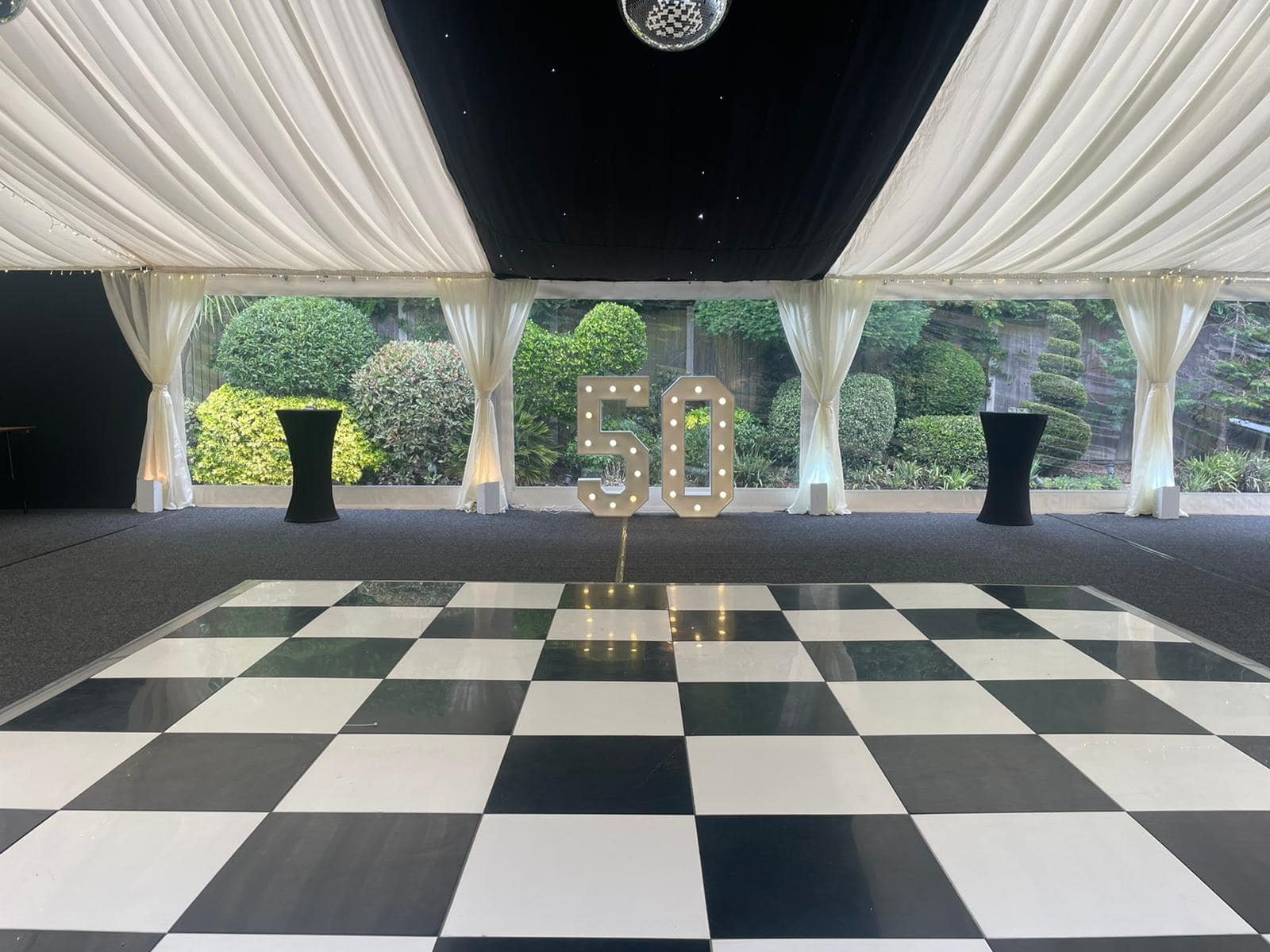 Decorated event space with a black and white checkered dance floor, white draped ceiling, large window with green bushes outside, illuminated '50' sign in the center, and two black high-top tables on either side.
