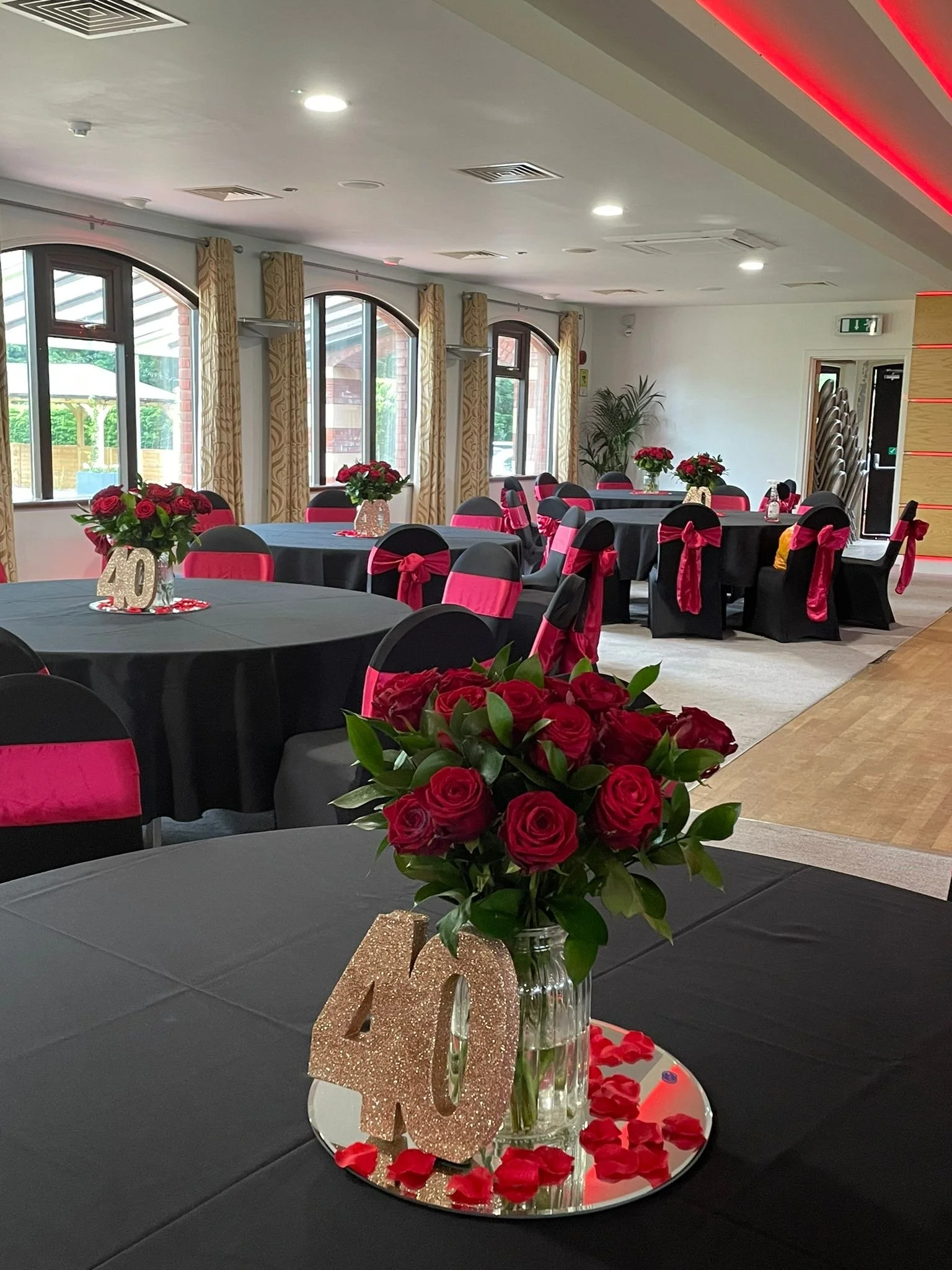 Decorated event hall with round tables covered in black tablecloths, featuring pink sashes on black chairs, red roses in vases, and a "40" glittery number centerpiece, suggesting a 40th celebration.
