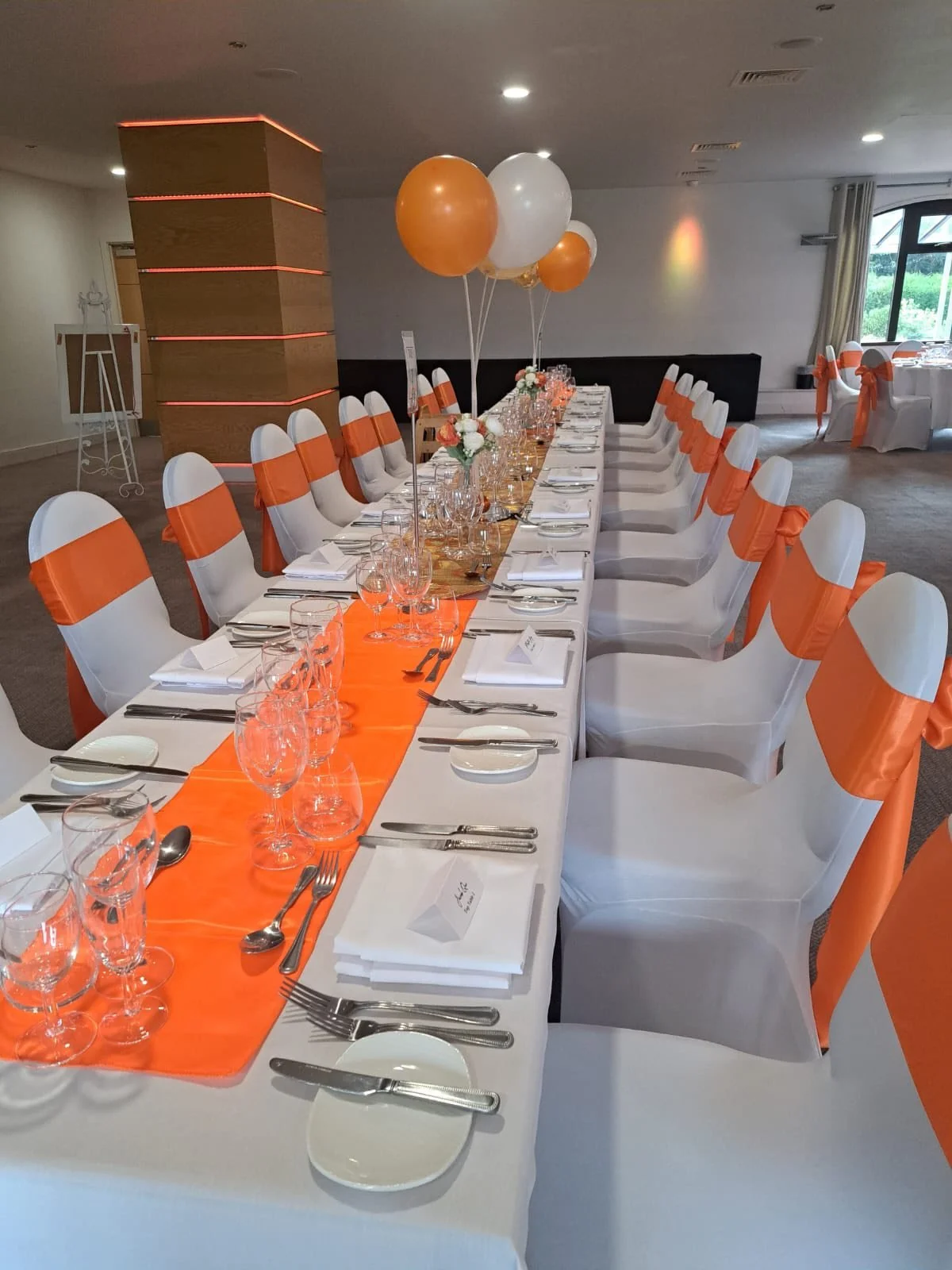 Long banquet table decorated with white tablecloth, orange runner, and white chairs with orange sashes. Centerpieces of balloons in orange, white, and gold, and small flower arrangements.
