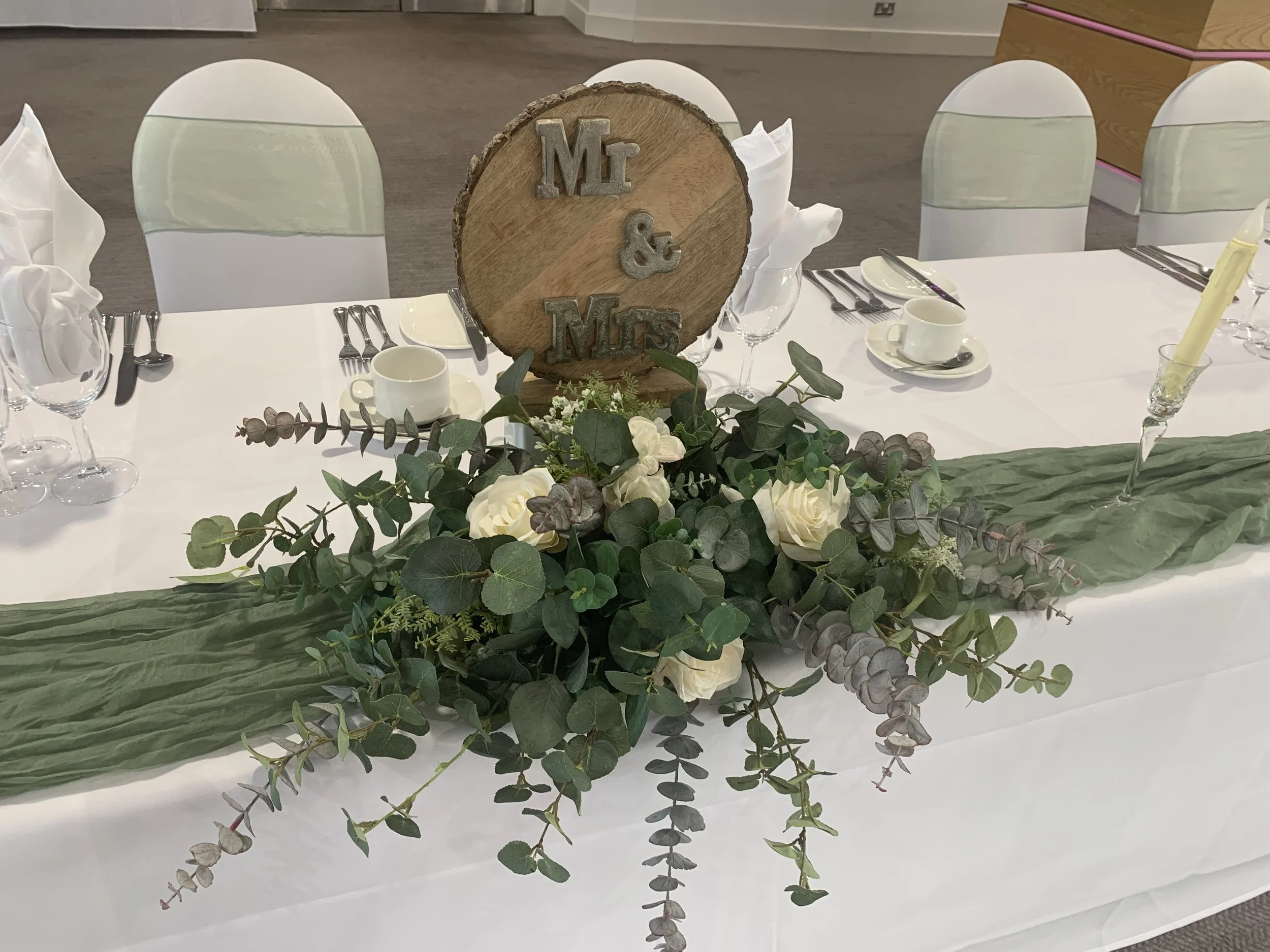A decorated wedding table with a floral centerpiece, a wooden round sign with initials 'M' and 'M' connected by an ampersand, set with place settings including cups, plates, and cutlery, and surrounded by white chairs with green sashes.