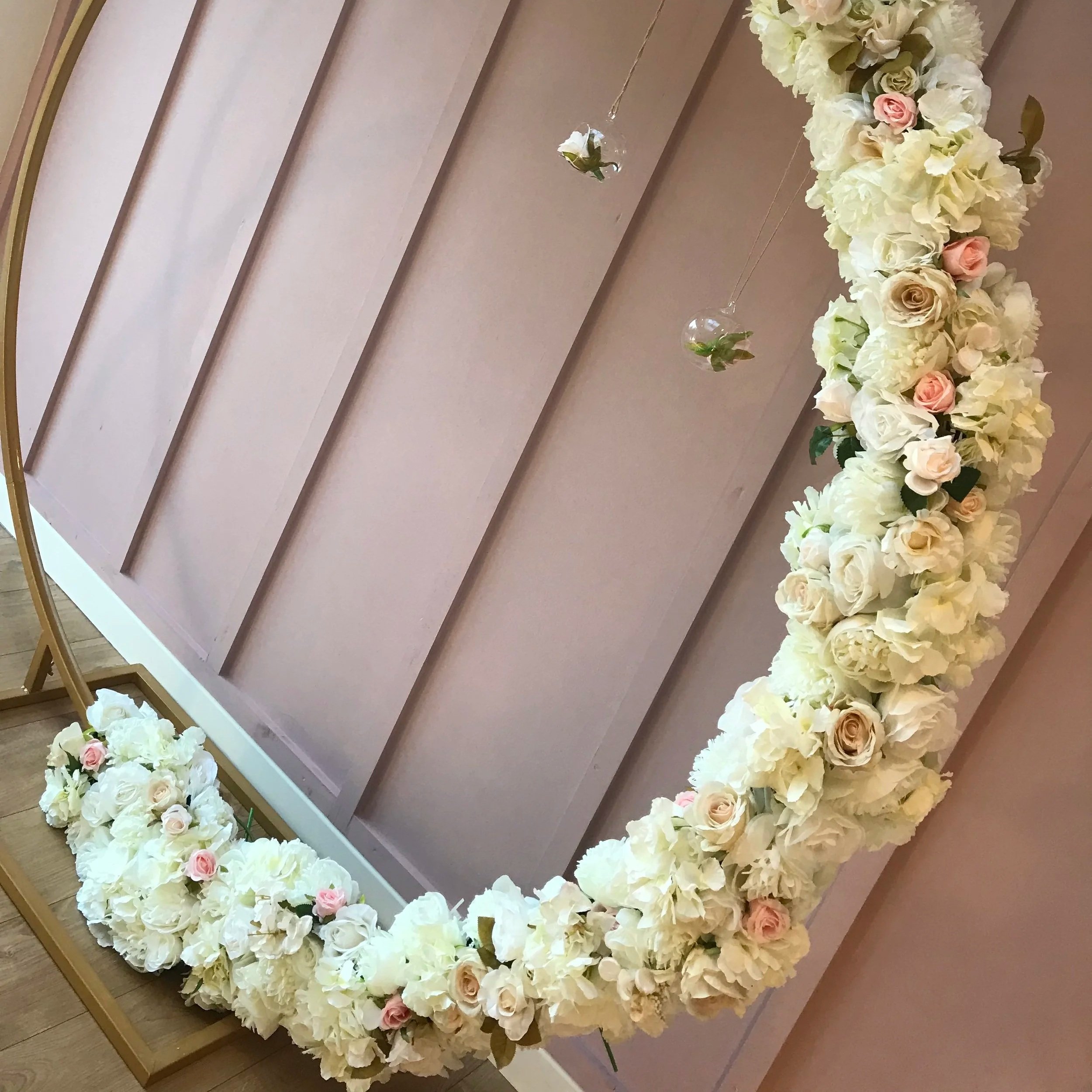 Round mirror decorated with white and pink roses and hydrangeas, with hanging glass orbs containing small flowers, on a pink wall.