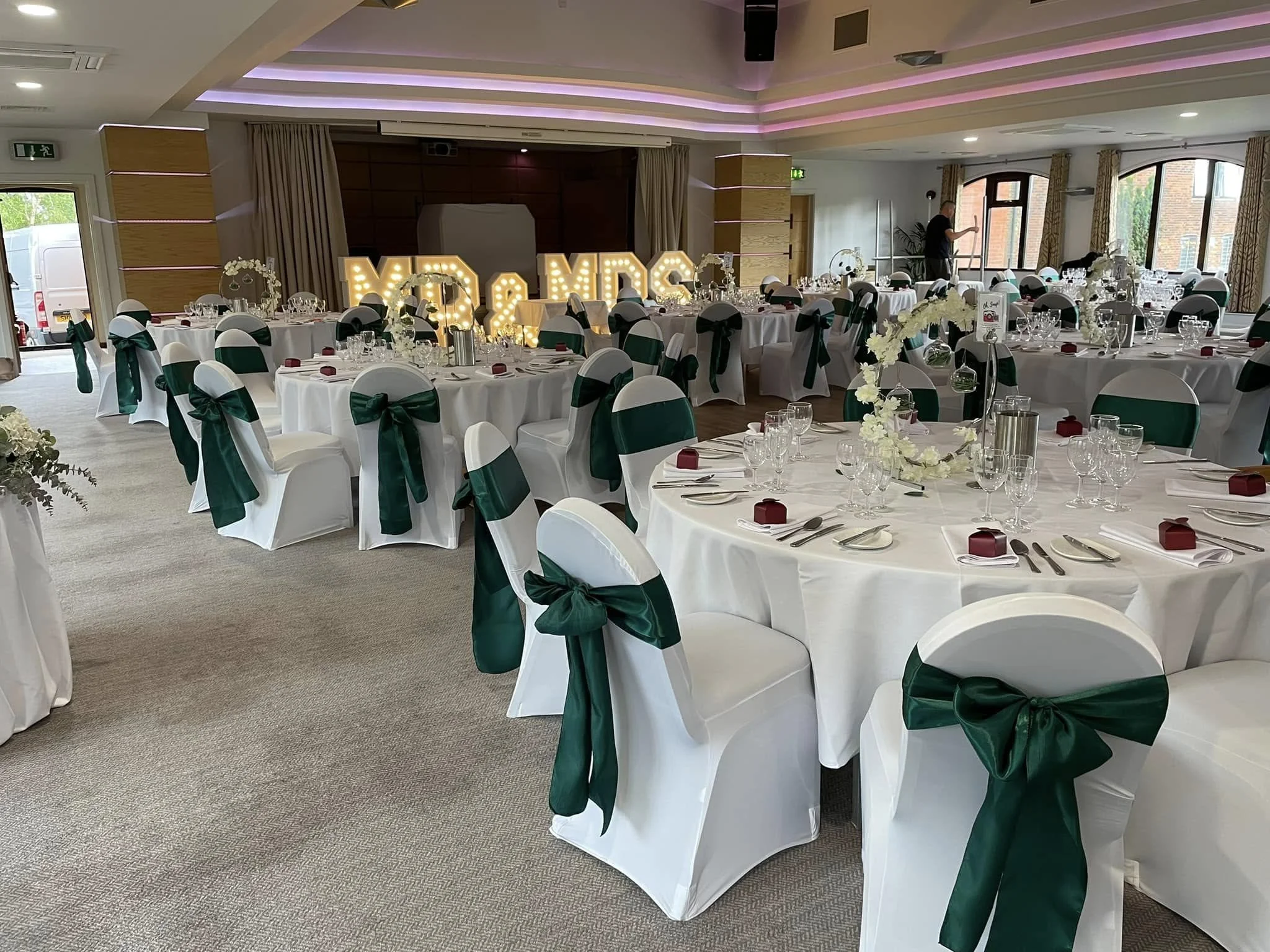 Wedding reception hall decorated with white tablecloths and chairs covered in white with green bows. Centerpieces include white flowers and candles. Large illuminated 'MR & MRS' sign in the background.