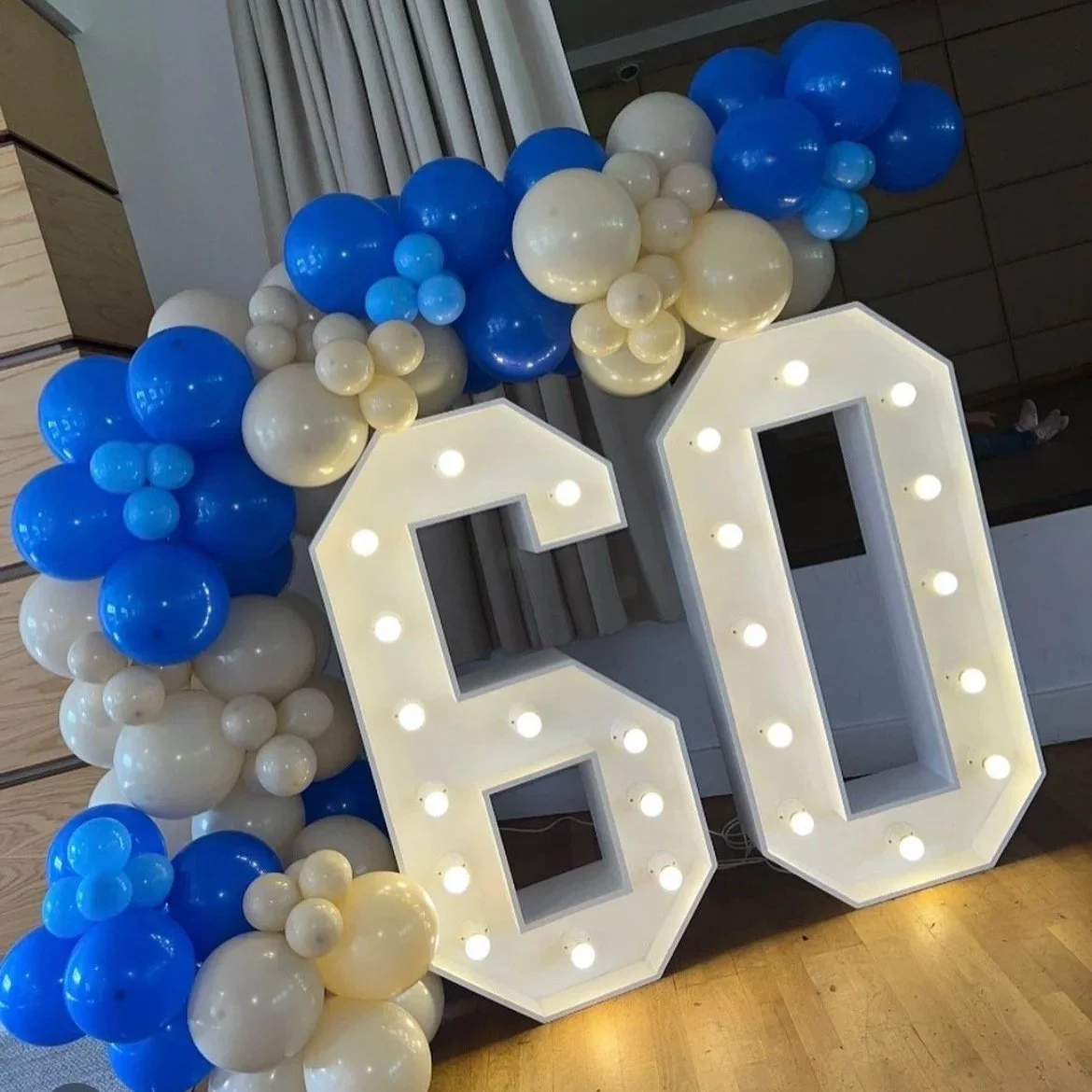 Large illuminated marquee numbers '60' decorated with white and blue balloons on a display stand.