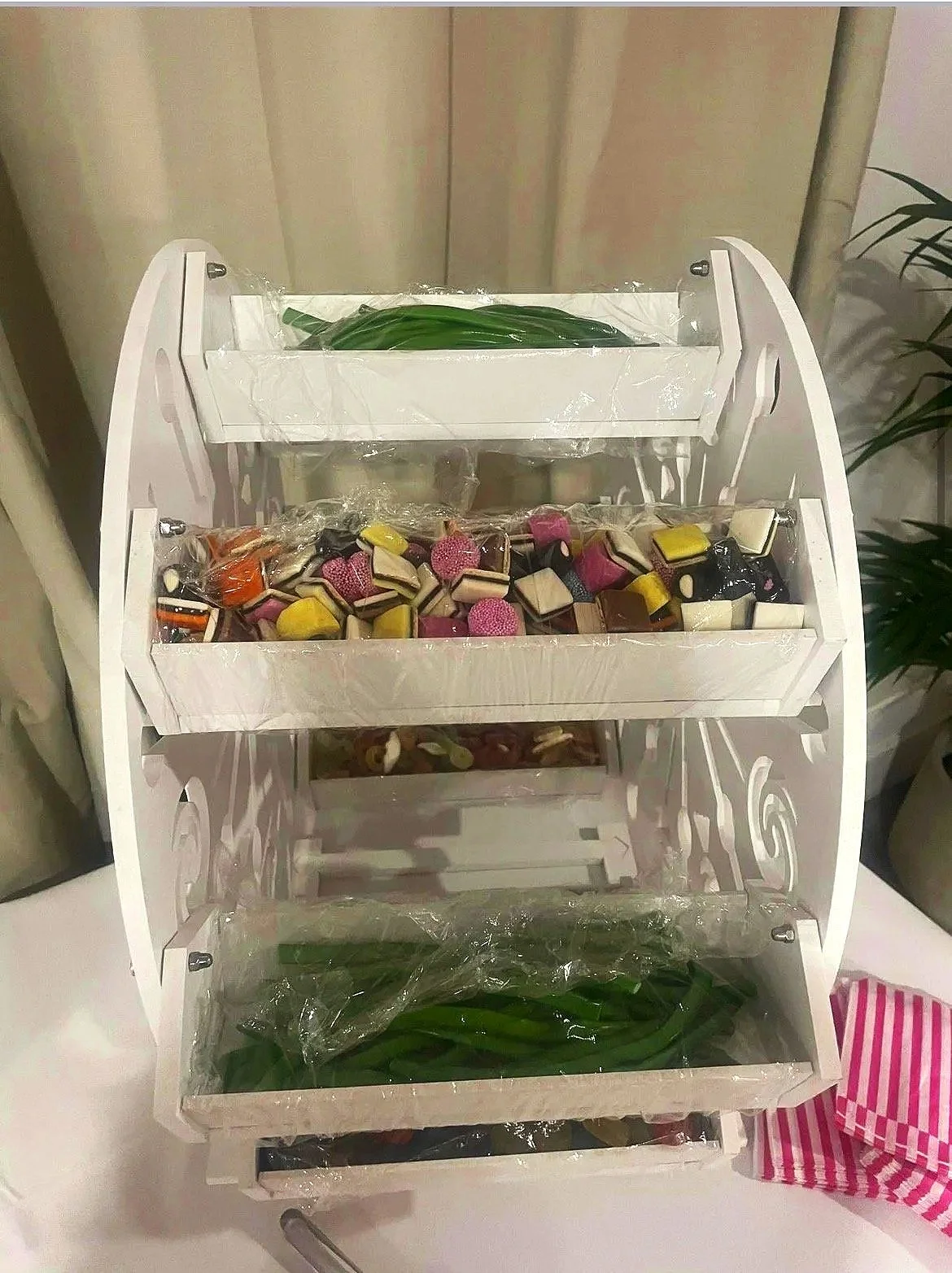 A white, tiered display stand with four shelves, each holding various wrapped candies and green leaves, situated on a table.