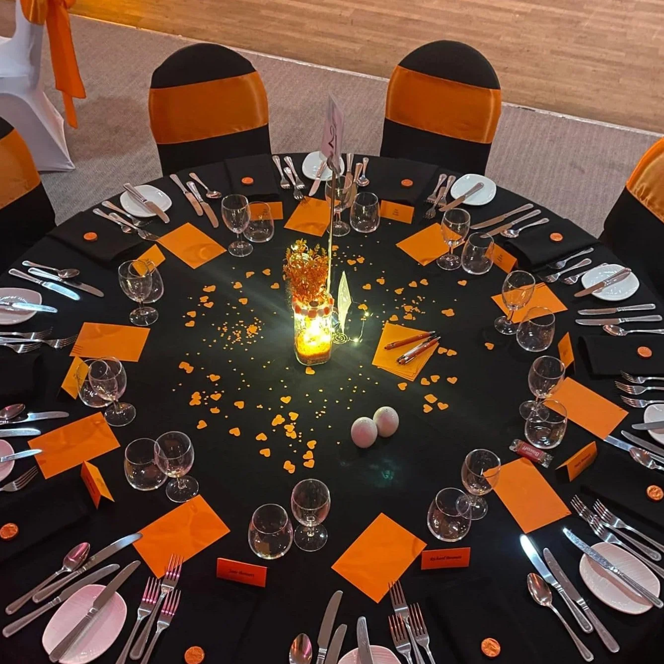 Round table decorated for an event with black tablecloth, orange accents, and heart-shaped confetti, set with plates, glasses, silverware, and orange place cards, centered around a lit decorative centerpiece.