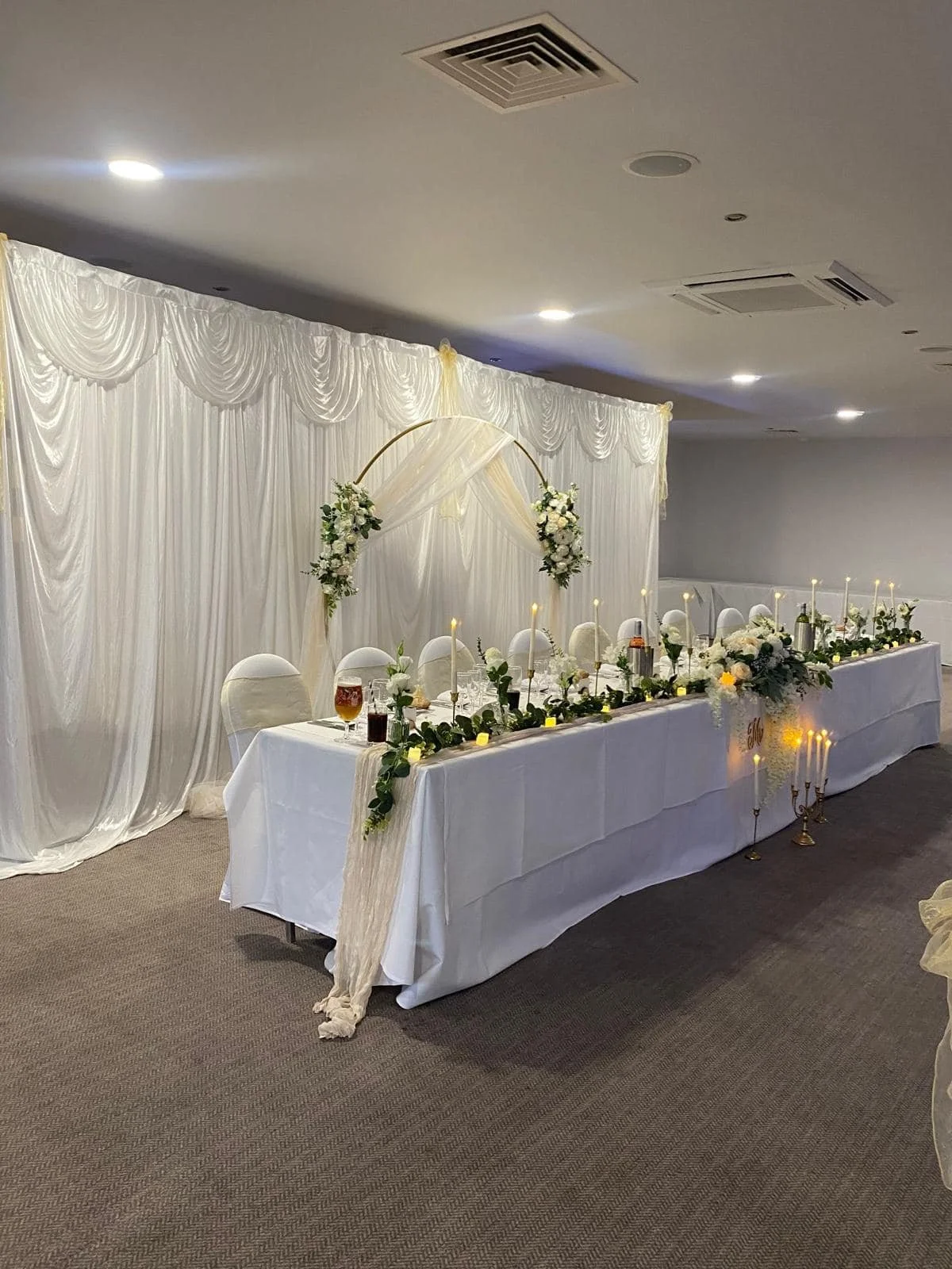Elegant wedding reception table decorated with white flowers, candles, and greenery, set against a white curtain backdrop with draped fabric and floral arrangements.