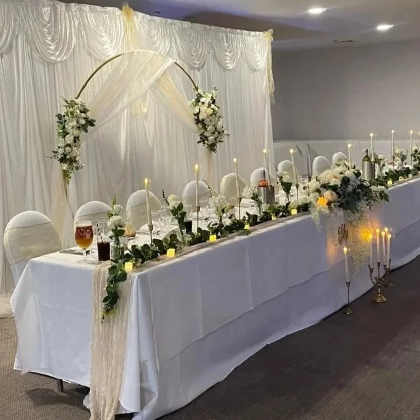 Elegant wedding reception table decorated with white flowers, candles, and greenery, with a white draped backdrop and floral arch.