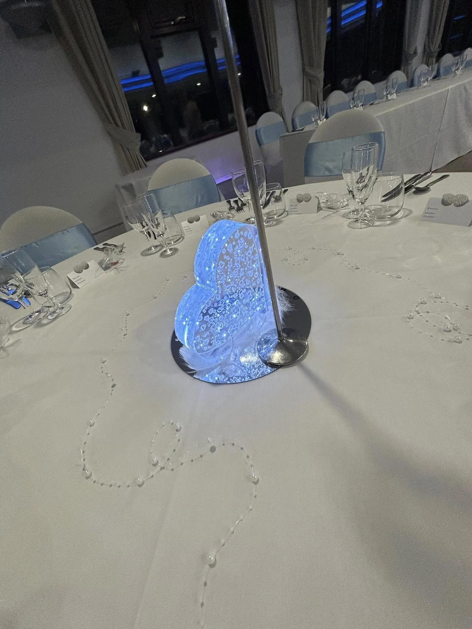 A decorated banquet table with an ice sculpture centerpiece, surrounded by white chairs with light blue sashes, glassware, and place cards.