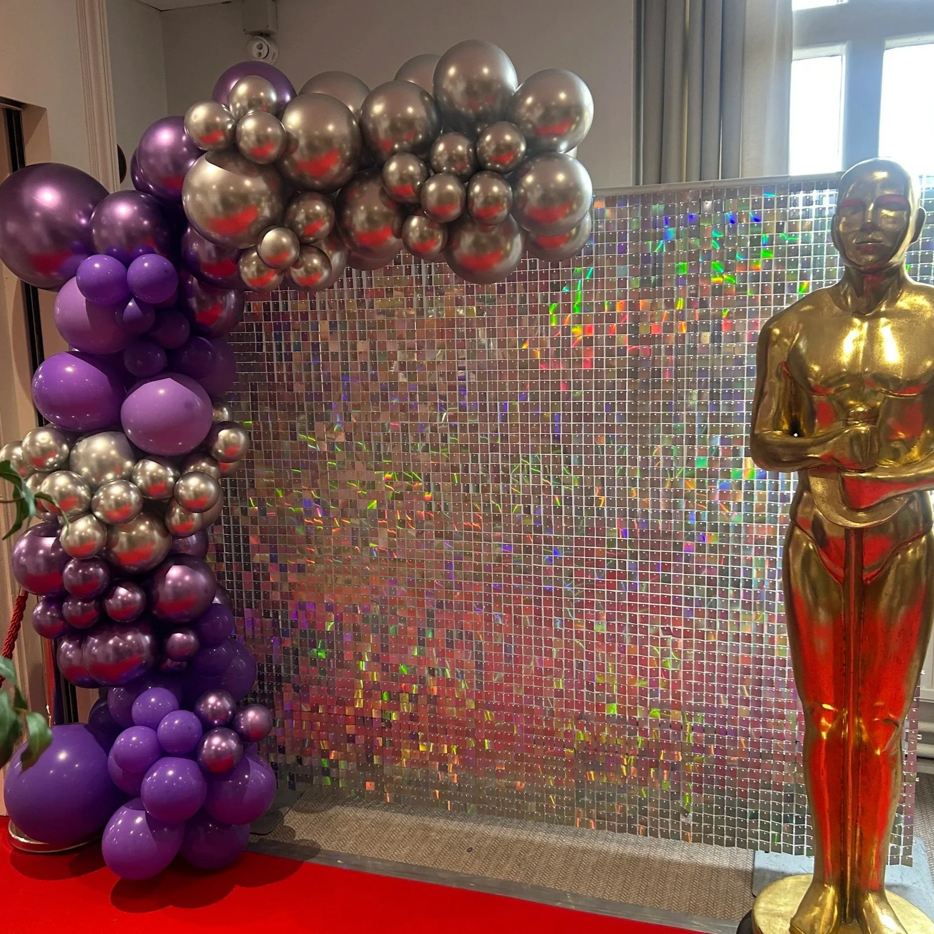 Decorative backdrop with purple and silver balloons, holographic mosaic wall, and a gold statue of a man.
