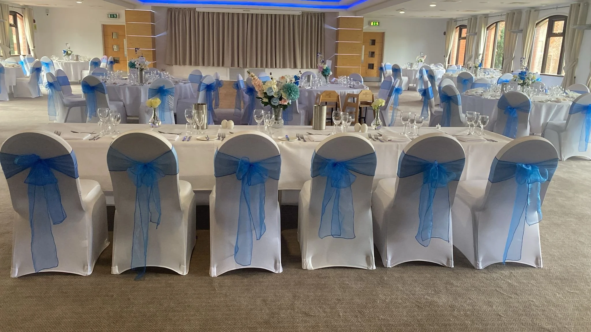 Wedding reception venue with round and long tables, decorated with blue and white flowers, white tablecloths, and white chairs adorned with blue ribbons, set with glassware and silverware.