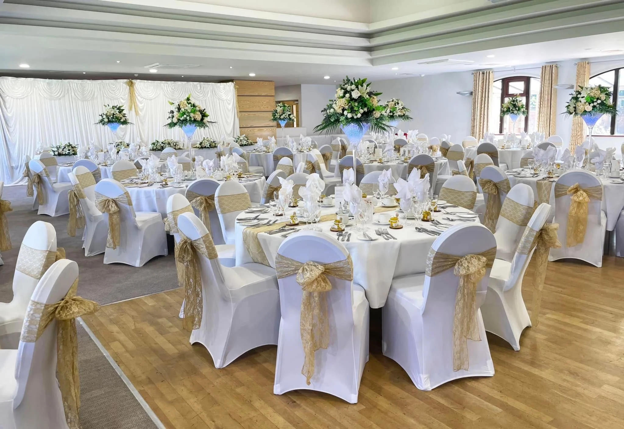 Elegant banquet hall decorated for a wedding reception with tables covered in white linens, gold sashes on the chairs, and large floral centerpieces with white flowers and greenery.