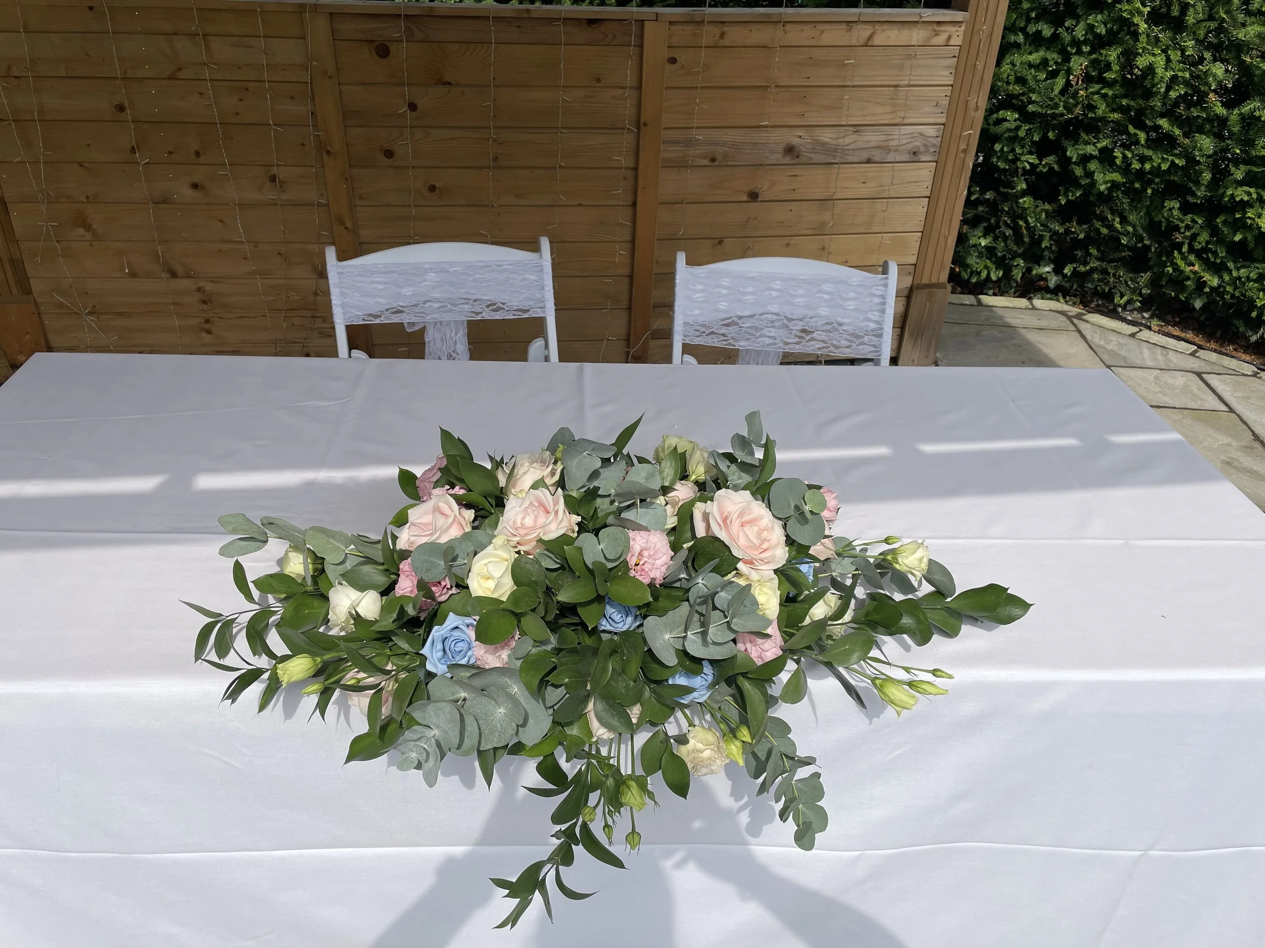 A table covered with a white tablecloth, decorated with a floral arrangement of pink, blue, and white flowers with green leaves, set outdoors with a wooden fence and green hedges in the background.
