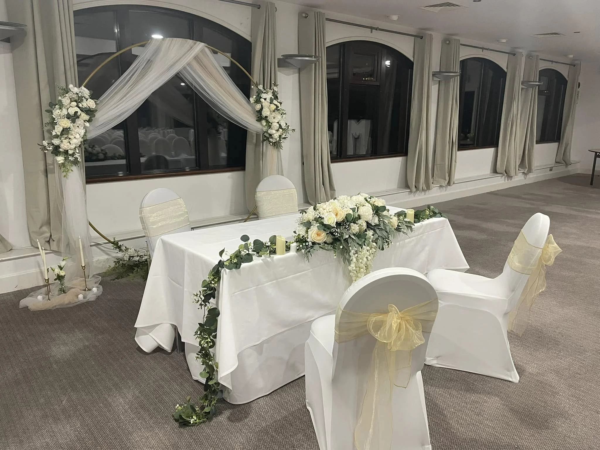 Wedding reception setup with a floral arch, decorated chairs, and a draped table with floral centerpiece in a banquet hall.