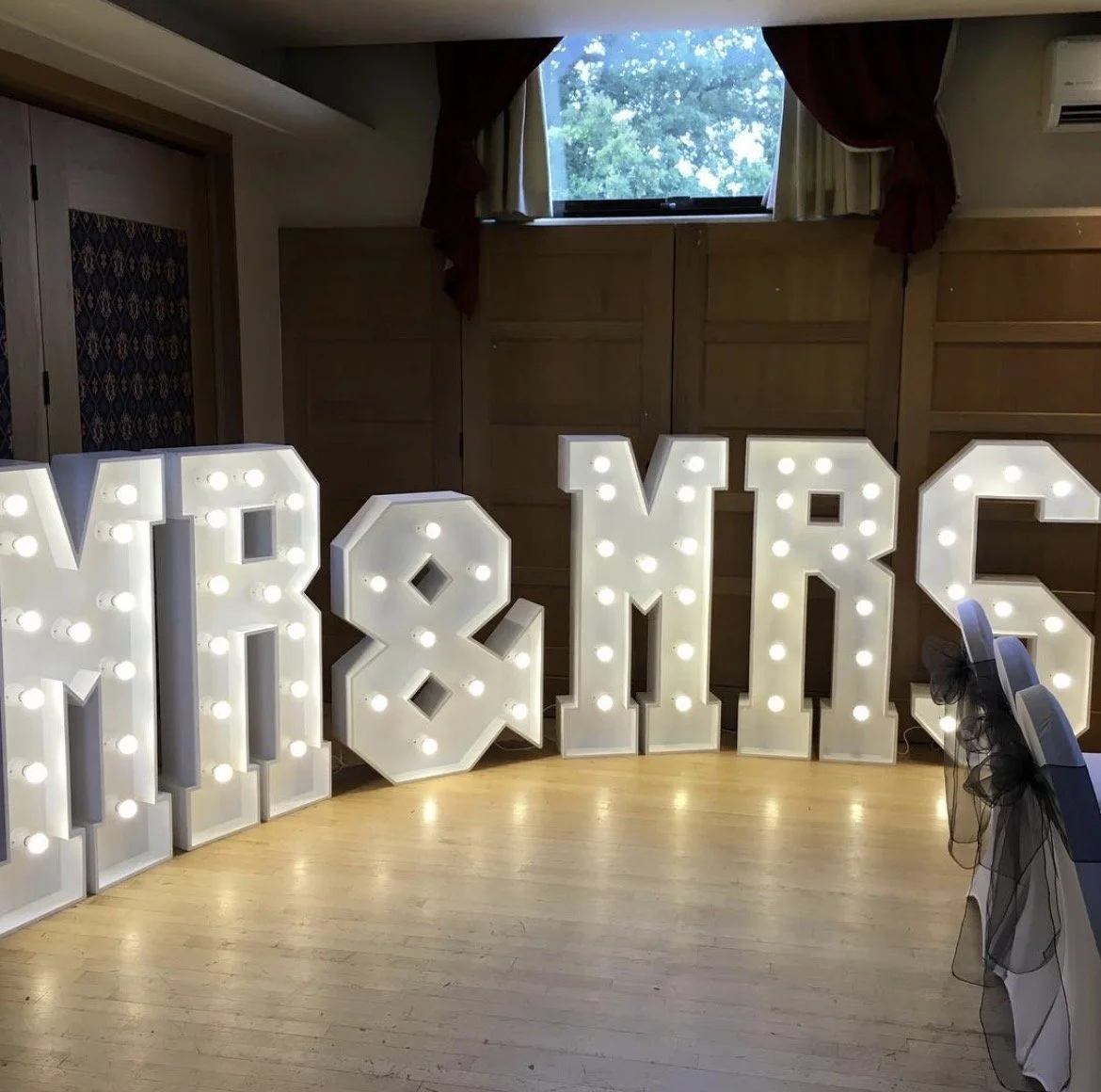 Large white marquee letters spelling 'MR & MRS' with light bulbs inside, set on a wooden floor in a decorated event space.