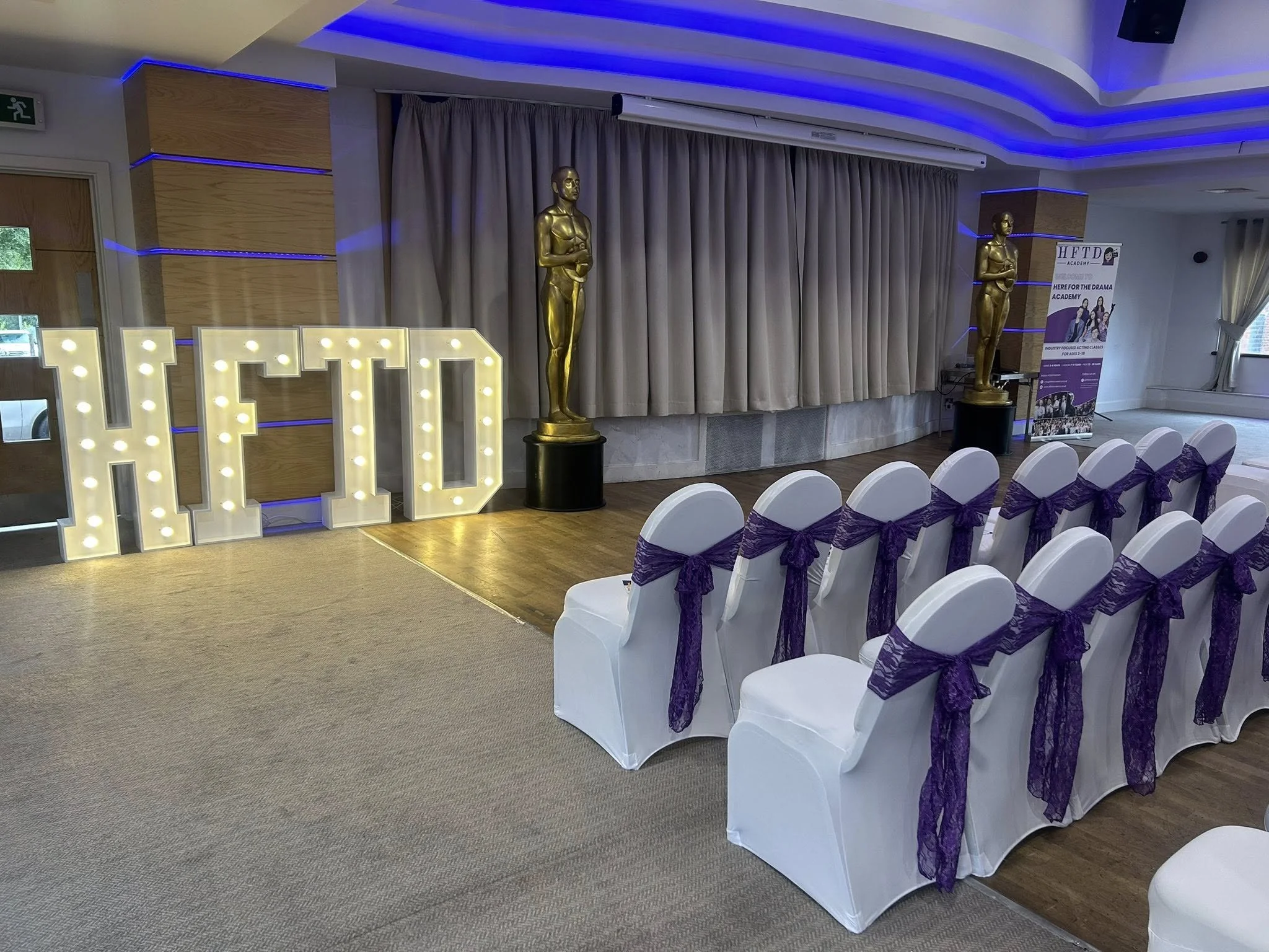An event space with a large lit sign spelling "HFD", two golden statue awards, rows of white chairs with purple sashes, and a purple and white promotional banner in the background.