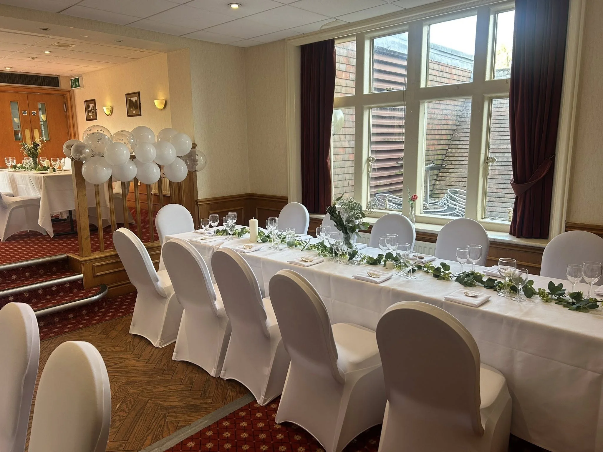 Decorated banquet hall with a long white table set with glassware, candles, and greenery, near a large window with burgundy curtains, and a cluster of white balloons on a wooden stand near the entrance.