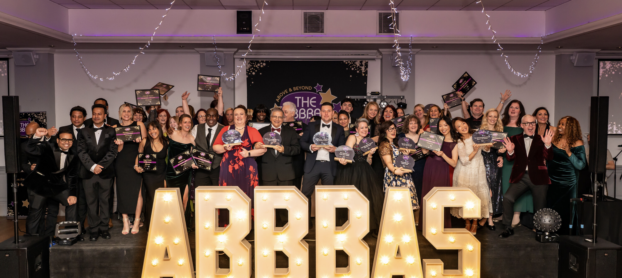 Group of people on stage at an awards event, holding plaques and certificates, with large illuminated letters spelling ABBAS in front, and festive decorations hanging from the ceiling.