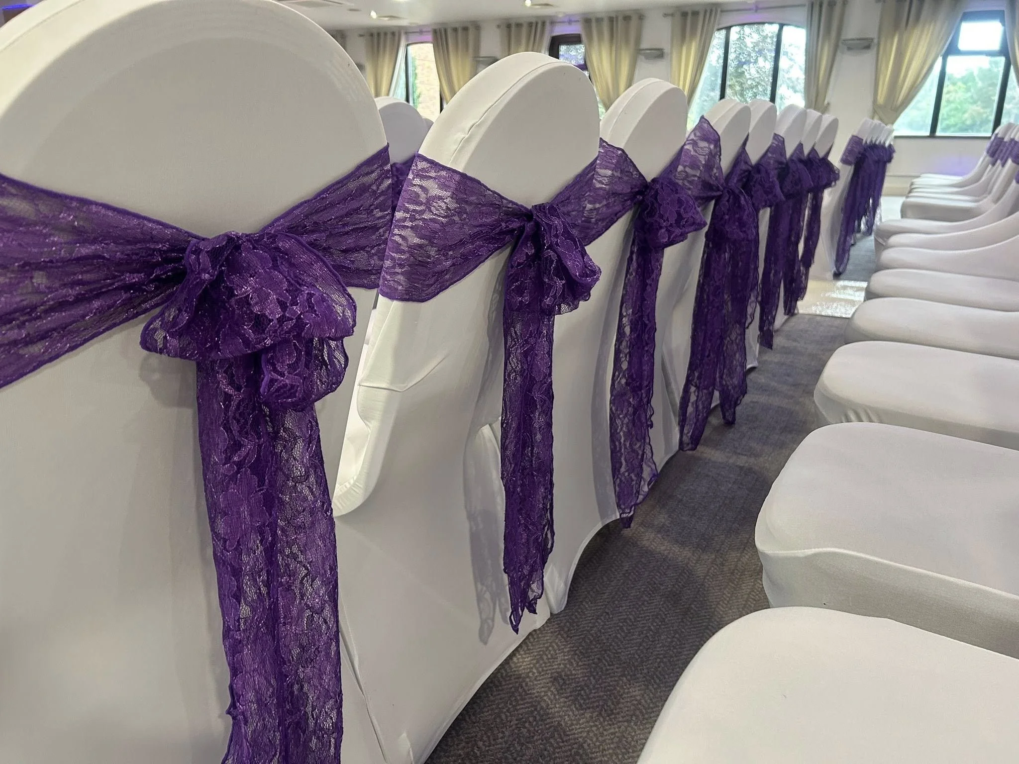 White chairs decorated with purple lace sashes tied in bows, arranged in a row in a decorated event venue.