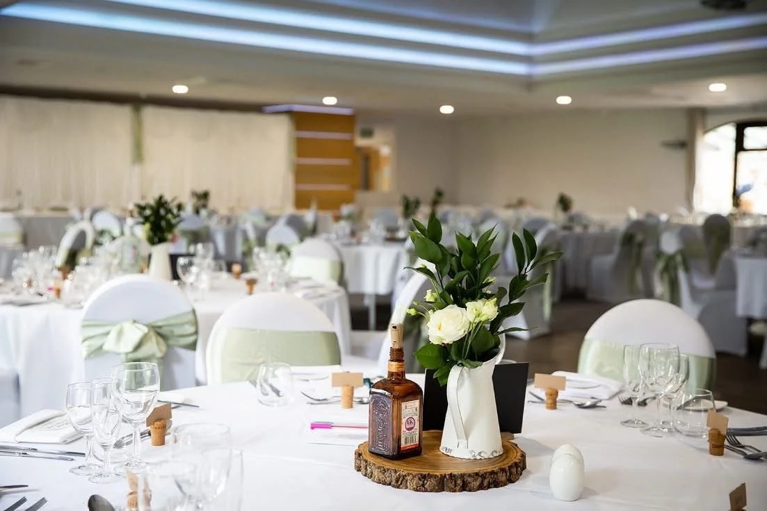 Round banquet table decorated for a formal event with a floral centerpiece, a bottle, and glassware in an elegant banquet hall